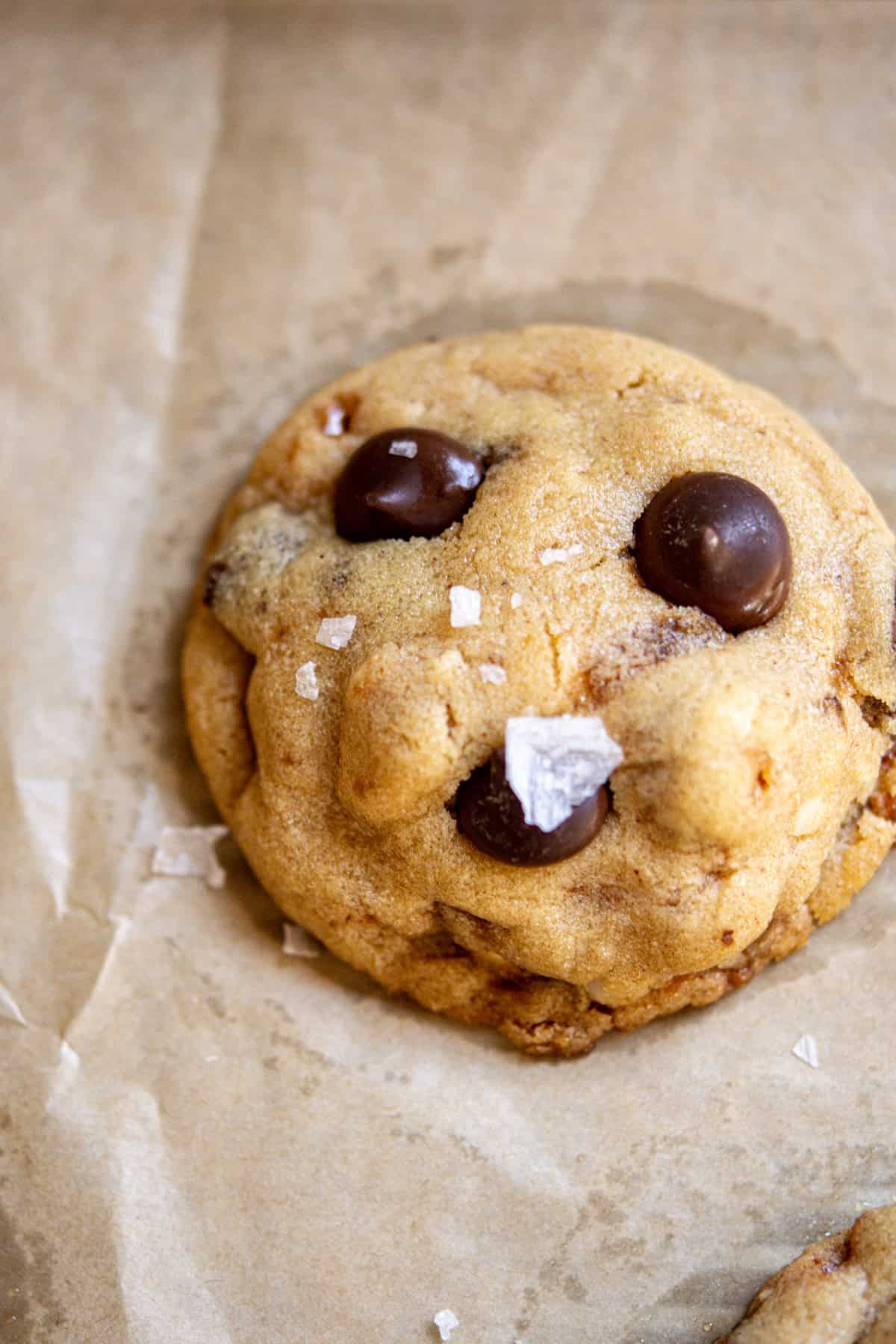 A chocolate chip cookie decorated to resemble a smiling face, with chocolate chips for eyes and mouth, and sprinkled with flaky sea salt, set on a sheet of parchment paper.