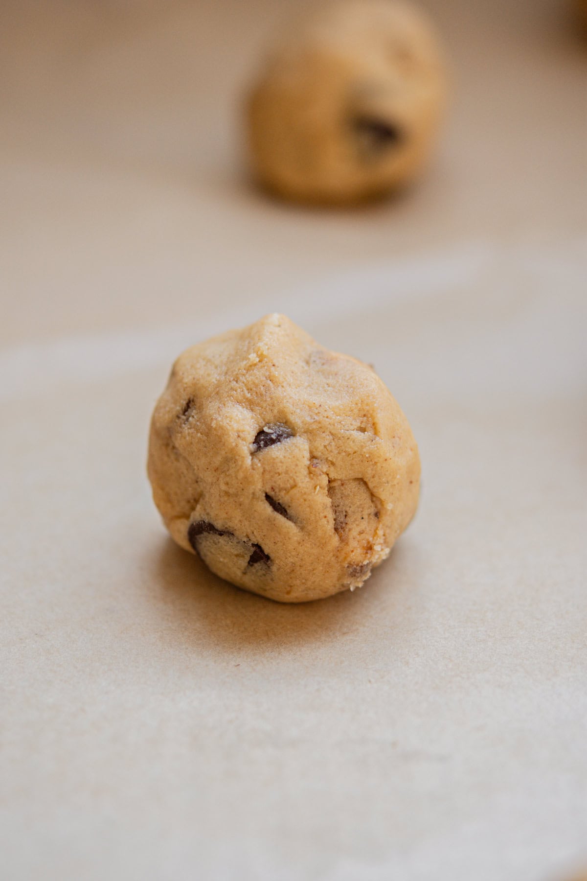A close-up of a raw chocolate hazelnut cookie dough ball on a sheet of parchment paper, with another dough ball blurred in the background.