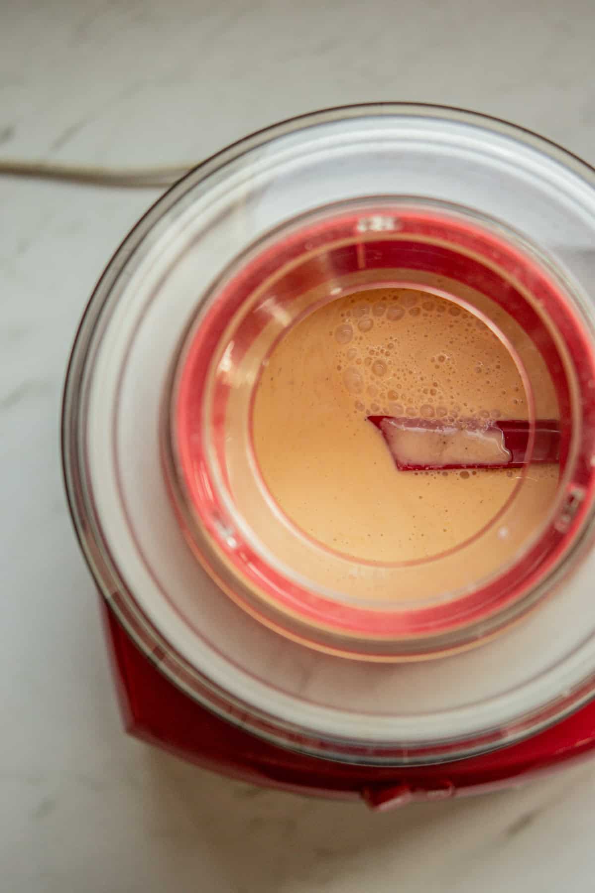 Top view of a red ice cream maker blending a light brown creamy mixture, with a red spatula stirring inside the transparent lid.