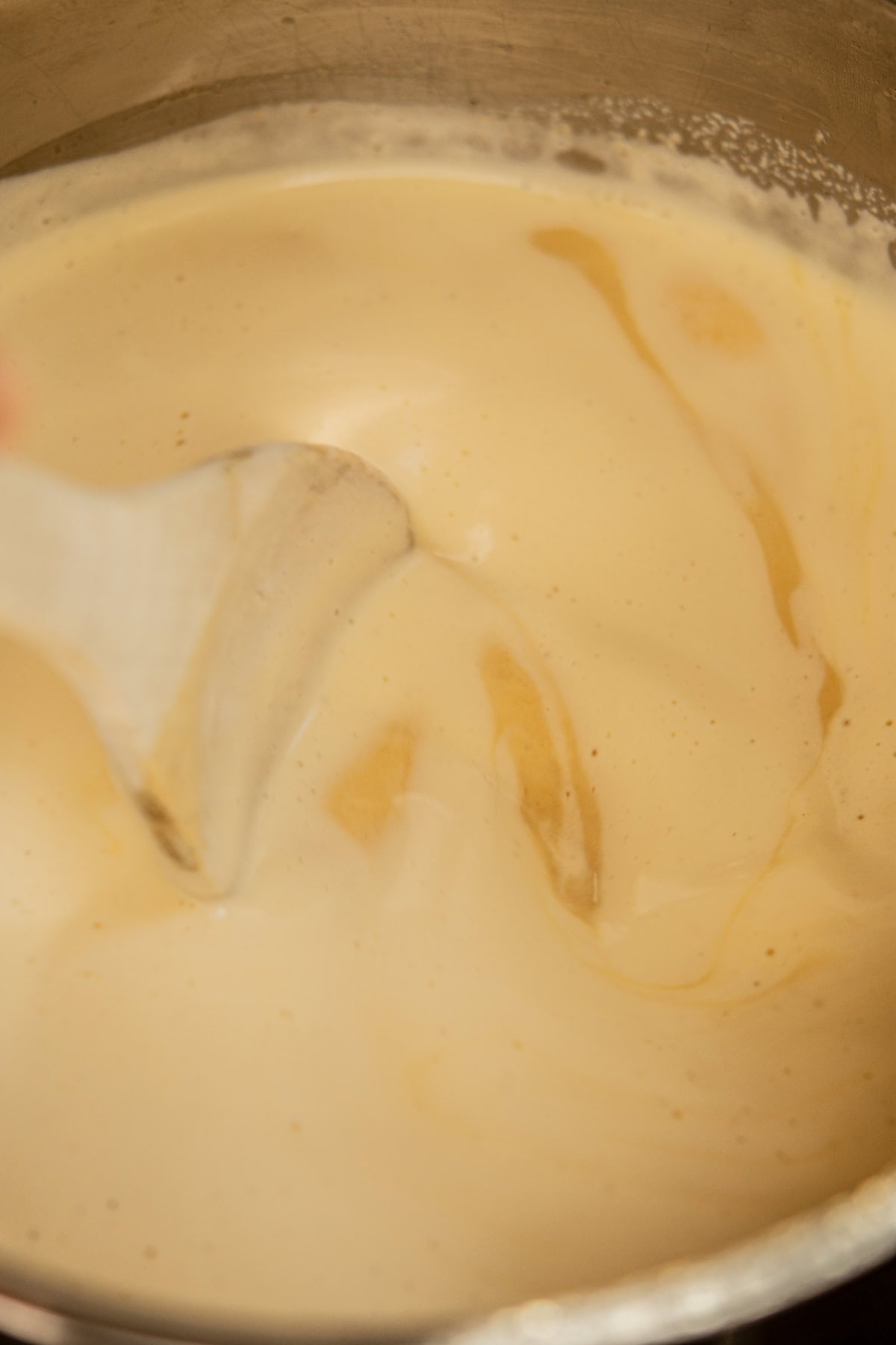 A close-up of a creamy, light beige mixture being stirred with a spatula in a metal bowl, showing smooth texture and swirls.