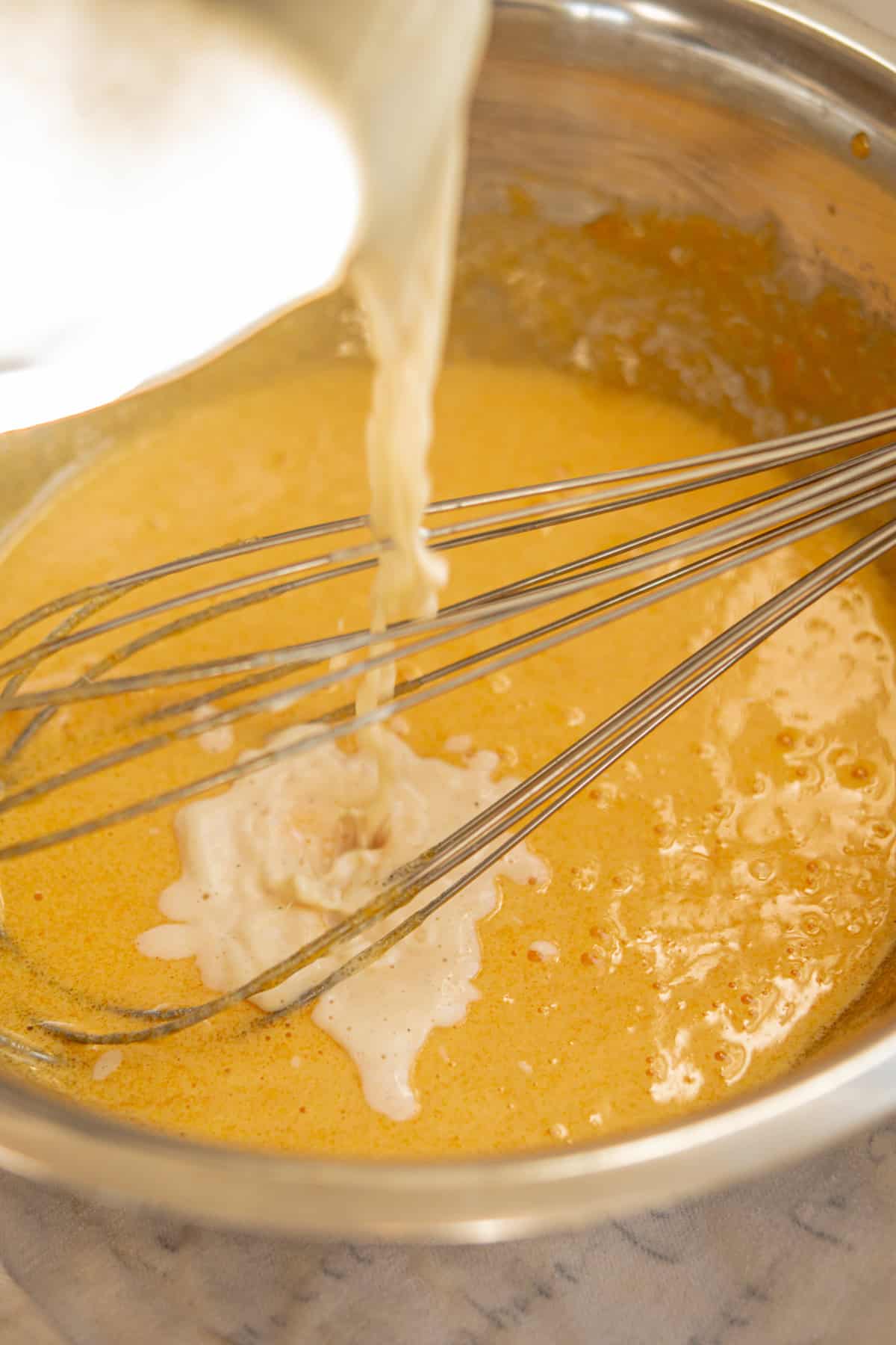 A close-up of a metal bowl with a yellow-orange batter being mixed by two whisks, while a creamy liquid is being poured into the bowl.