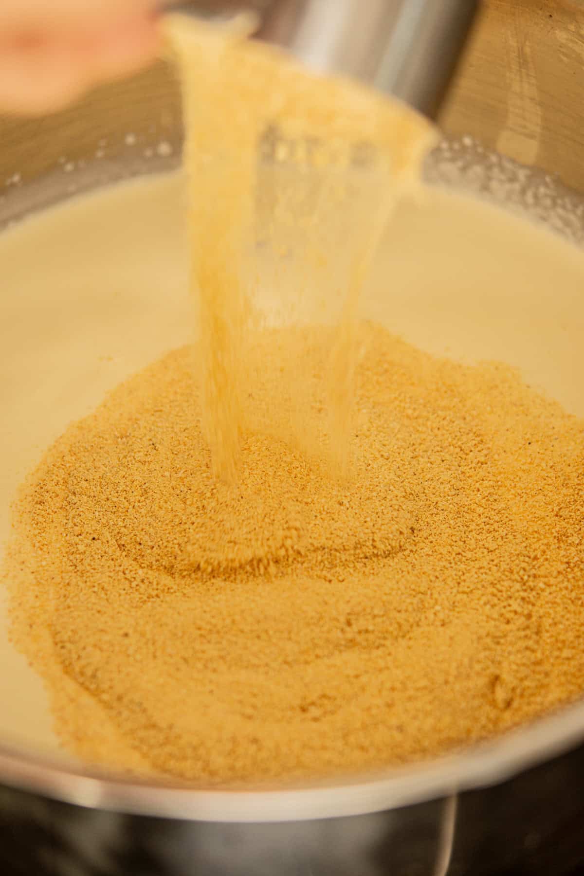 Breadcrumbs are being poured from a metal cup into a creamy mixture in a stainless steel bowl.