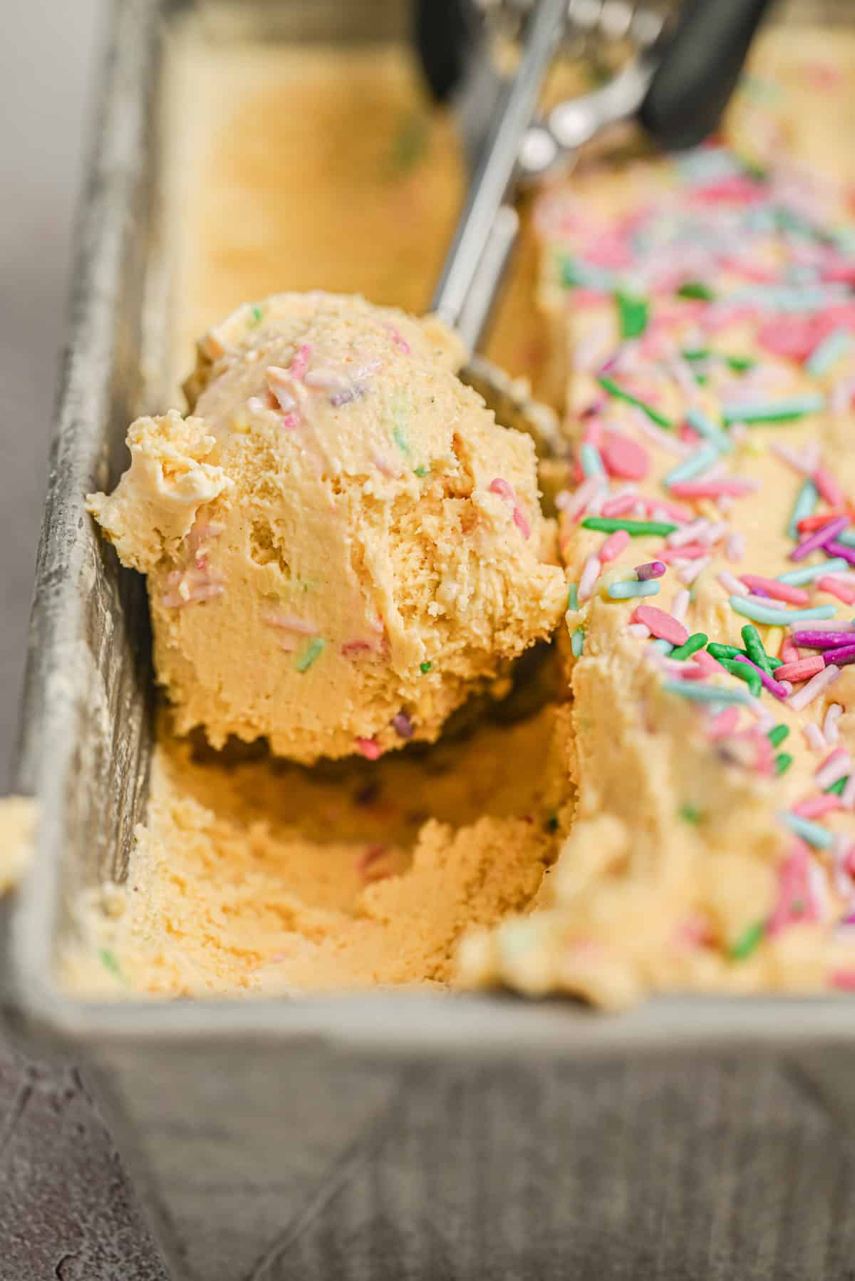 A close-up of a metal scoop lifting creamy yellow ice cream with colorful rainbow sprinkles from a metal container. The ice cream is soft and textured, with sprinkles scattered on top.