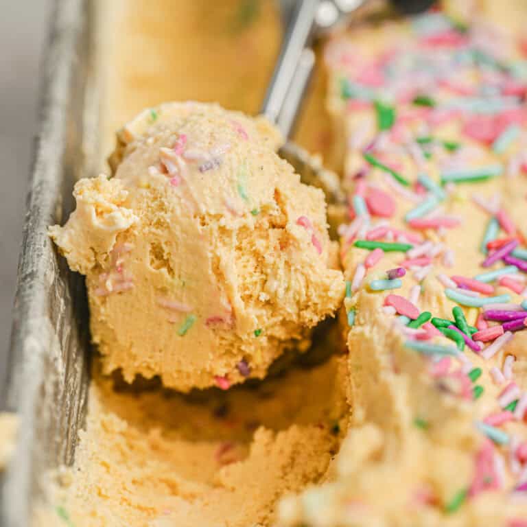 A close-up of a metal funfetti ice cream scoop holding a scoop of yellow ice cream with colorful sprinkles, next to more sprinkled ice cream in a loaf pan.