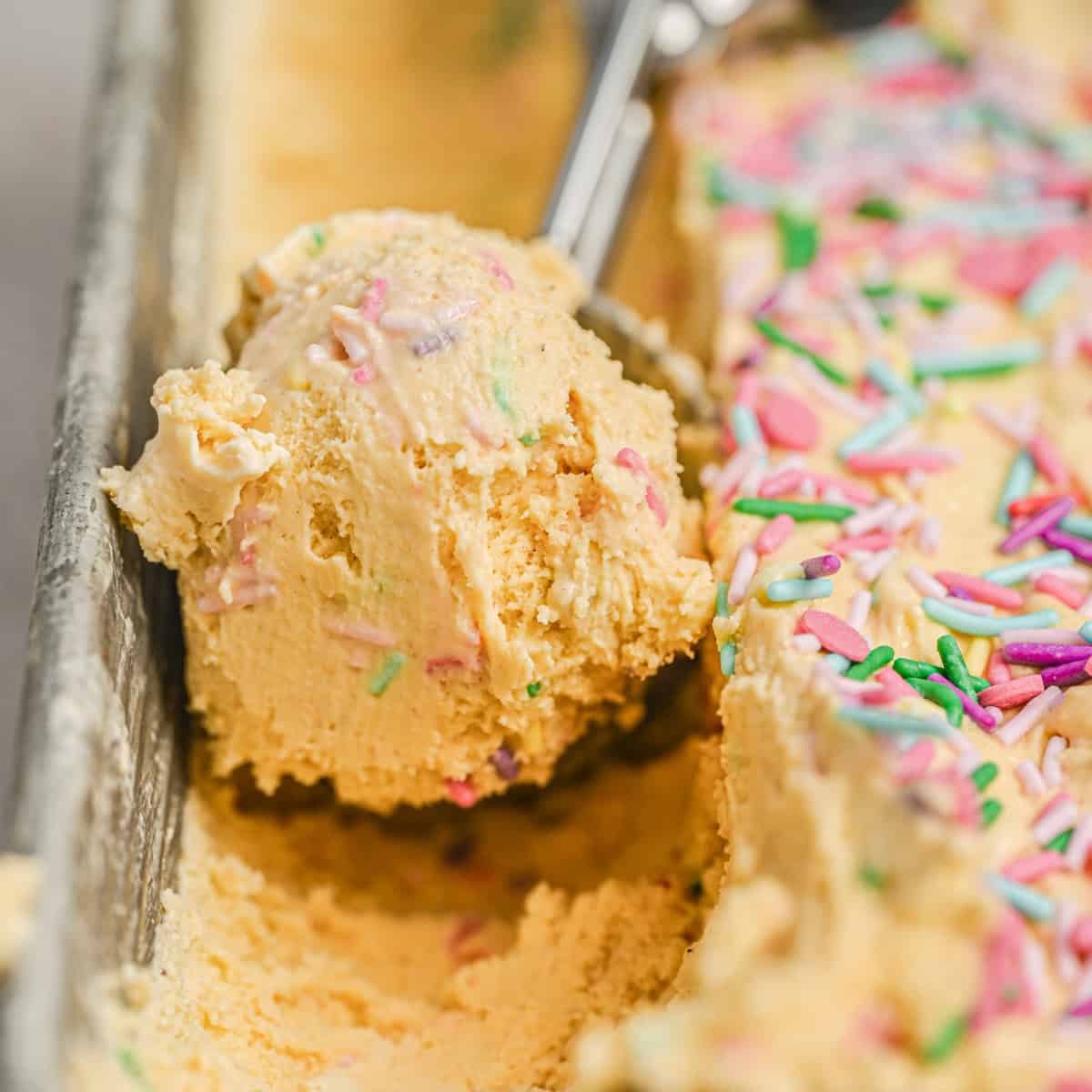 A close-up of a metal funfetti ice cream scoop holding a scoop of yellow ice cream with colorful sprinkles, next to more sprinkled ice cream in a loaf pan.