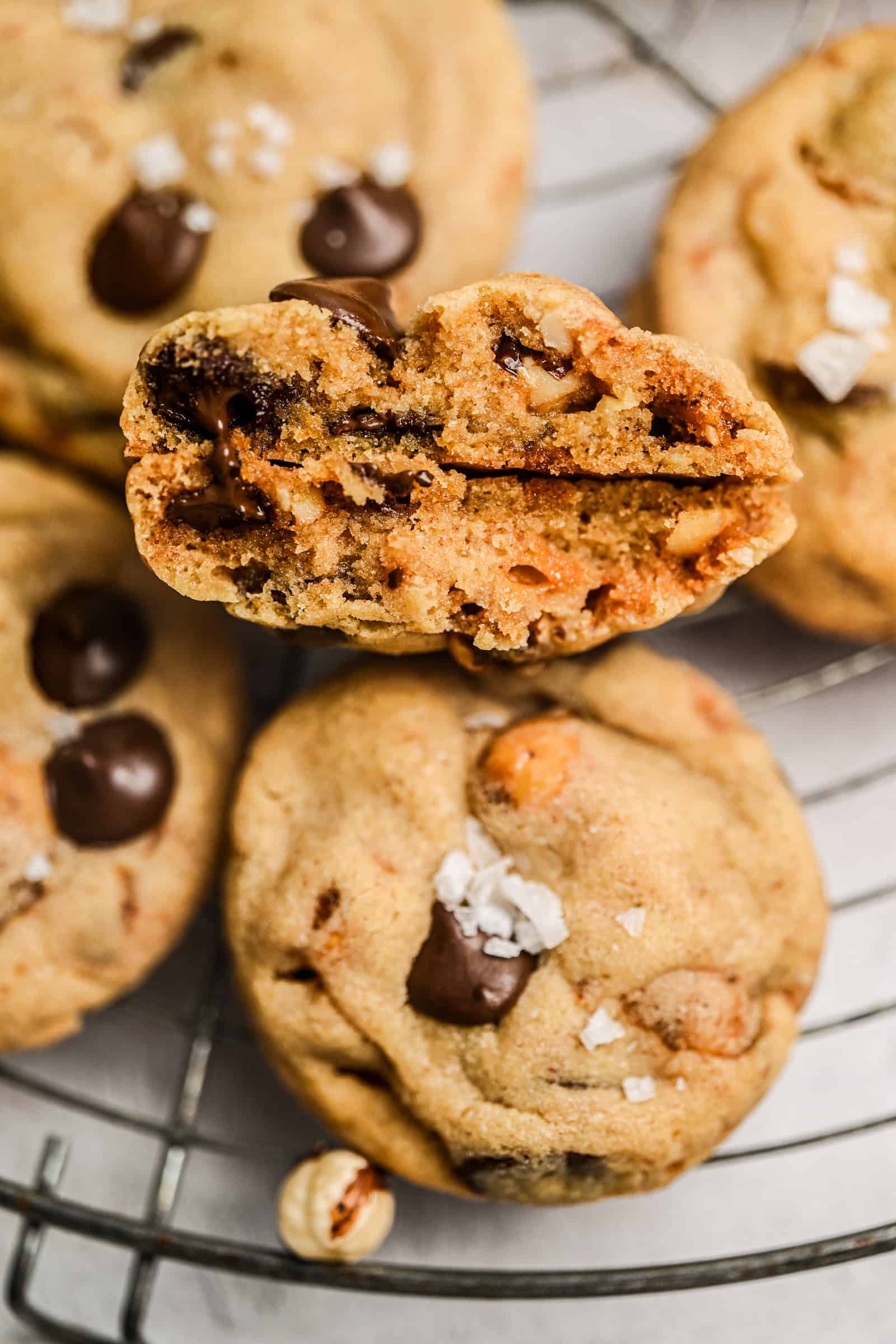 A close-up of hazelnut chocolate chip cookies on a wire rack, with one cookie broken in half to reveal a soft, gooey center. Some cookies are topped with flaky sea salt.