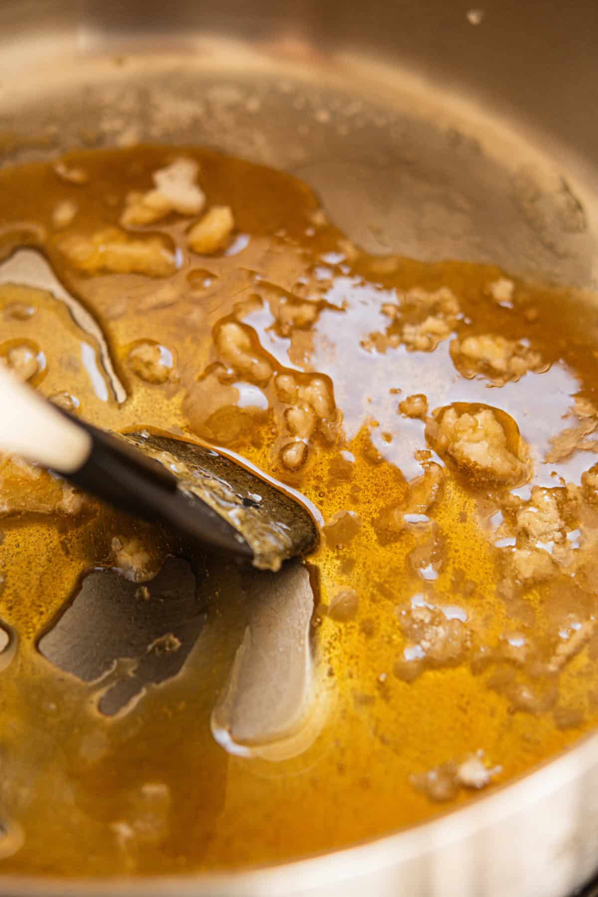 A close-up of a saucepan with melted brown sugar and butter being stirred with a black and white spatula. The mixture appears golden and bubbly.