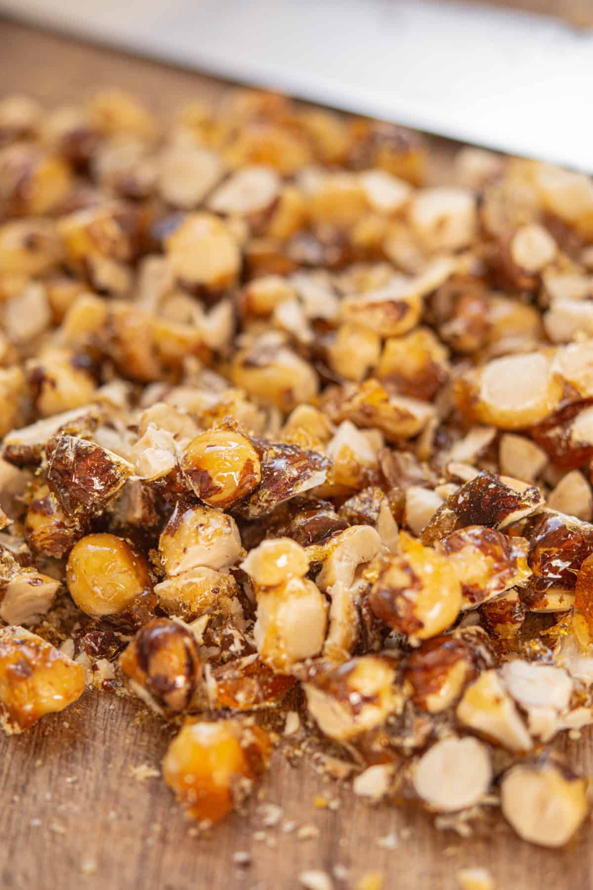 Close-up of chopped caramelized nuts on a wooden surface, with a blurred knife visible in the background. The nuts are shiny and golden brown, suggesting a crunchy texture.