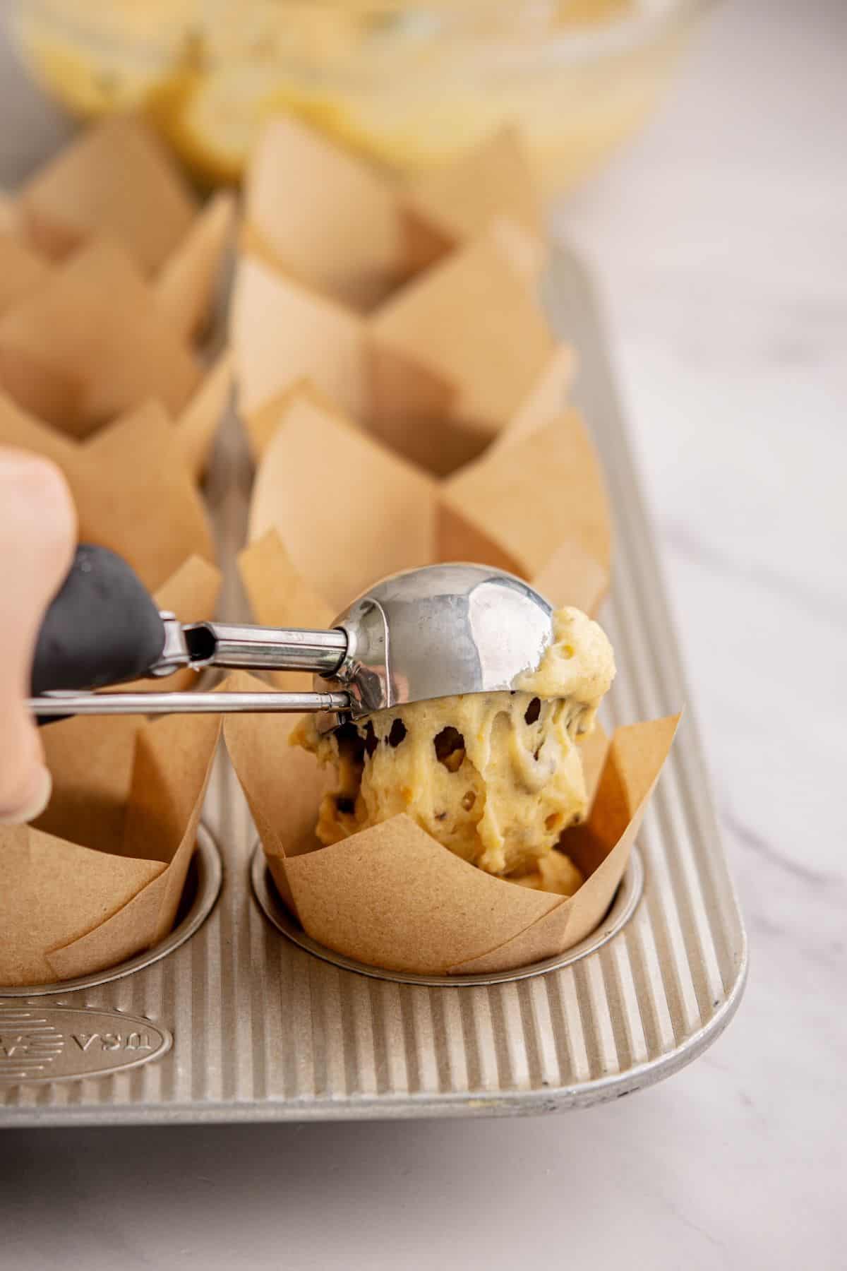 A hand uses an ice cream scoop to place chocolate chip muffin batter, inspired by Funfetti Ice Cream, into a brown paper liner in a muffin tin on a marble countertop.