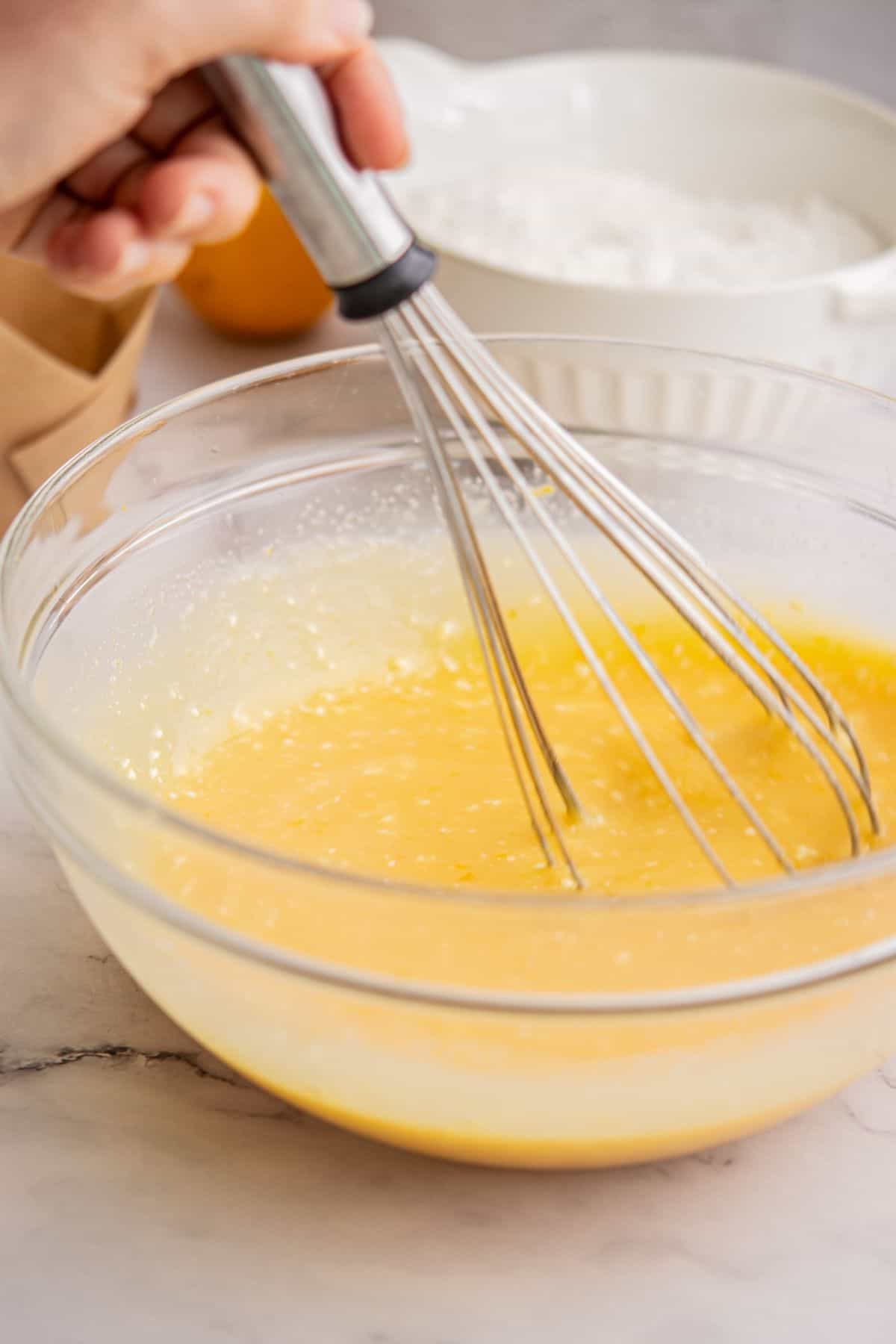 A hand holding a whisk is mixing a yellow Funfetti Ice Cream batter in a glass bowl, with a bowl of flour and a brown paper bag visible in the background on a marble countertop.