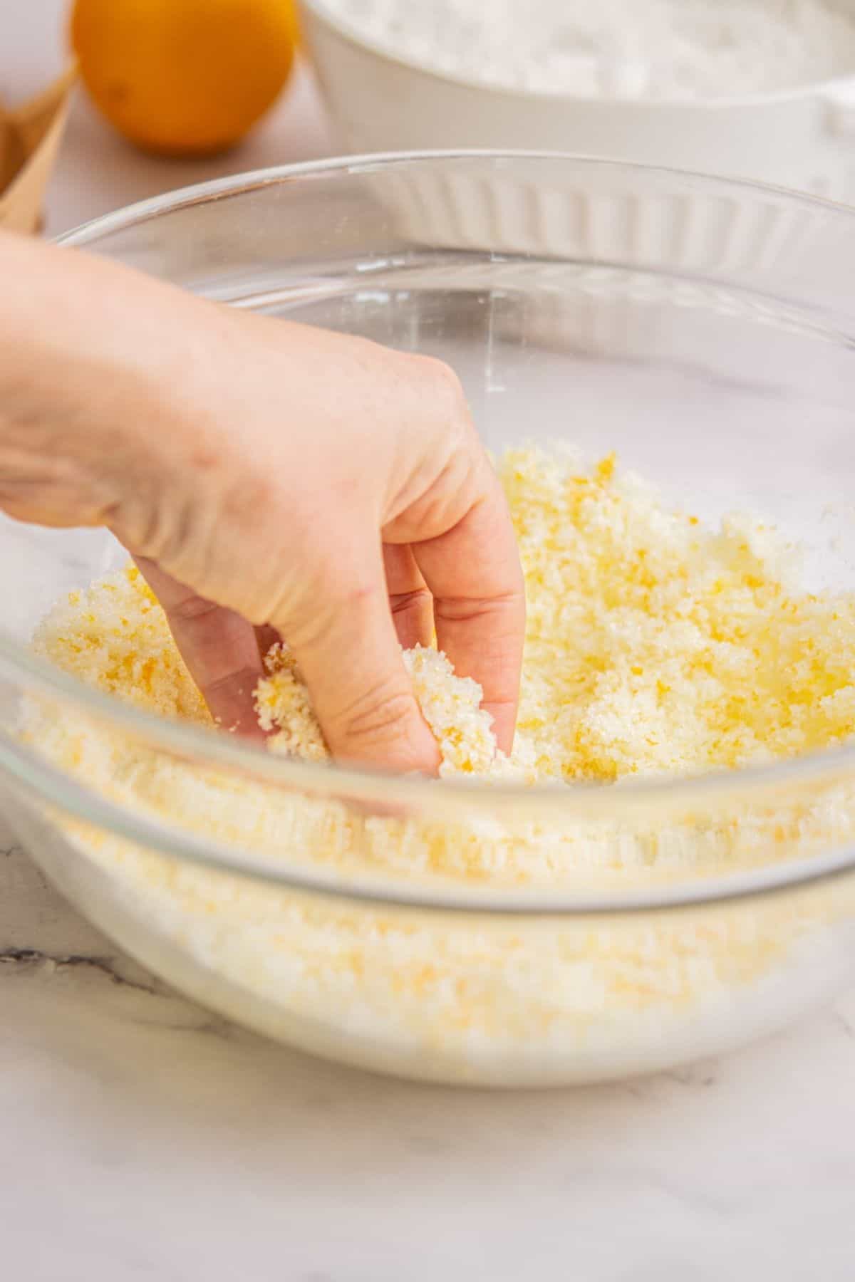A hand mixing or pinching flour and grated orange zest together in a clear glass bowl, preparing a base that pairs perfectly with Funfetti Ice Cream, with a white bowl and an orange in the background on a light-colored surface.