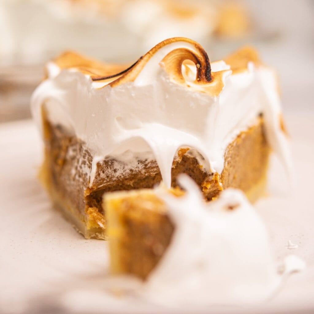 A close-up of a slice of pie with a golden-brown crust, creamy pumpkin filling, and a thick layer of toasted, fluffy meringue on top—reminiscent of a classic Strawberry Meringue Pie—with some meringue dripping down the sides.