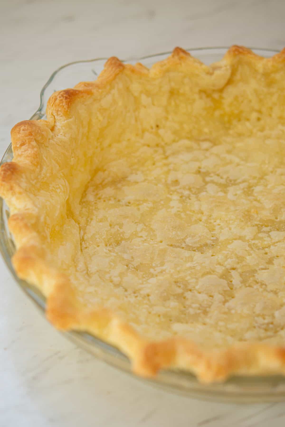 A close-up of a baked, empty pie crust with golden, flaky edges in a clear glass pie dish, set on a light-colored countertop.