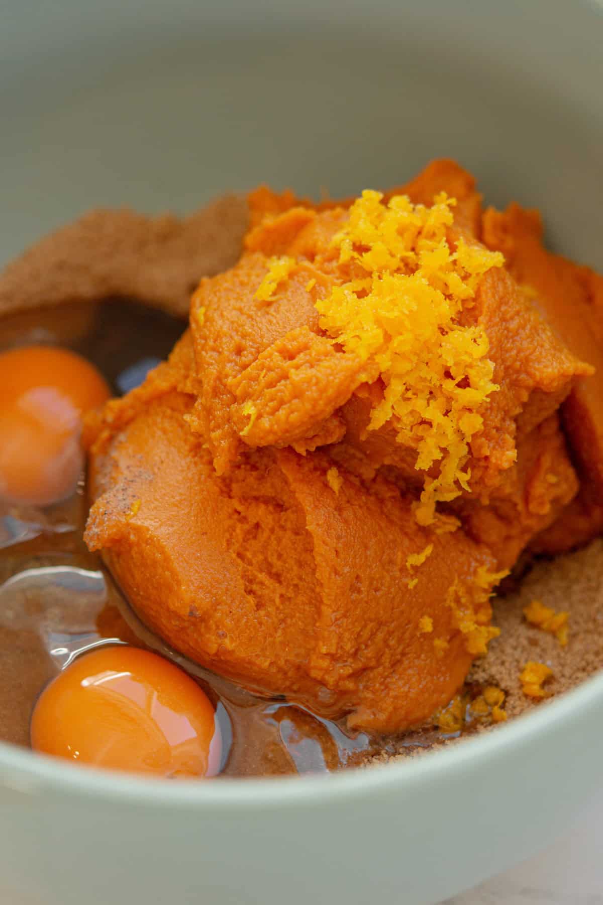 A mixing bowl containing raw eggs, brown sugar, pumpkin puree, and freshly grated orange zest, ready to be combined for baking.