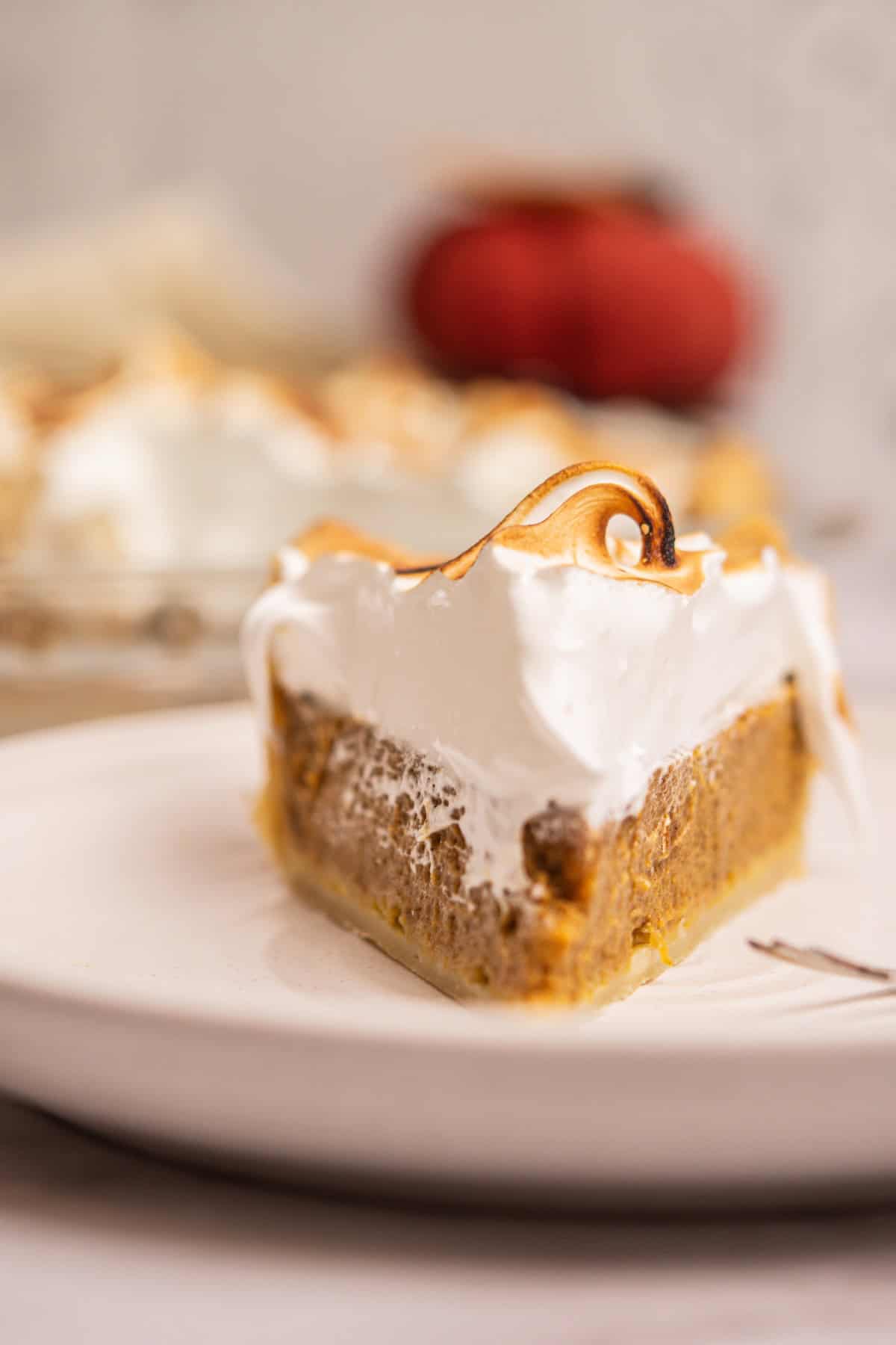 A close-up of a slice of pumpkin pie topped with toasted meringue on a white plate, with more pie and a pumpkin blurred in the background.