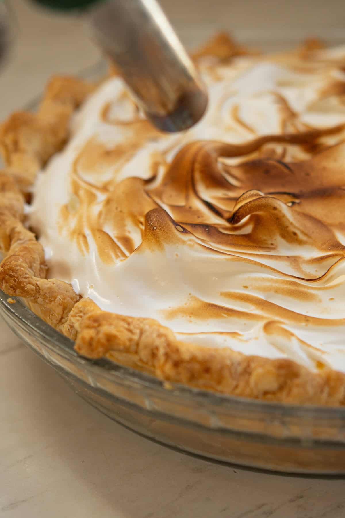 A close-up of a pie with golden-brown, toasted meringue on top. A kitchen torch is being used to brown the swirled meringue, which sits on a flaky, baked crust in a glass pie dish.