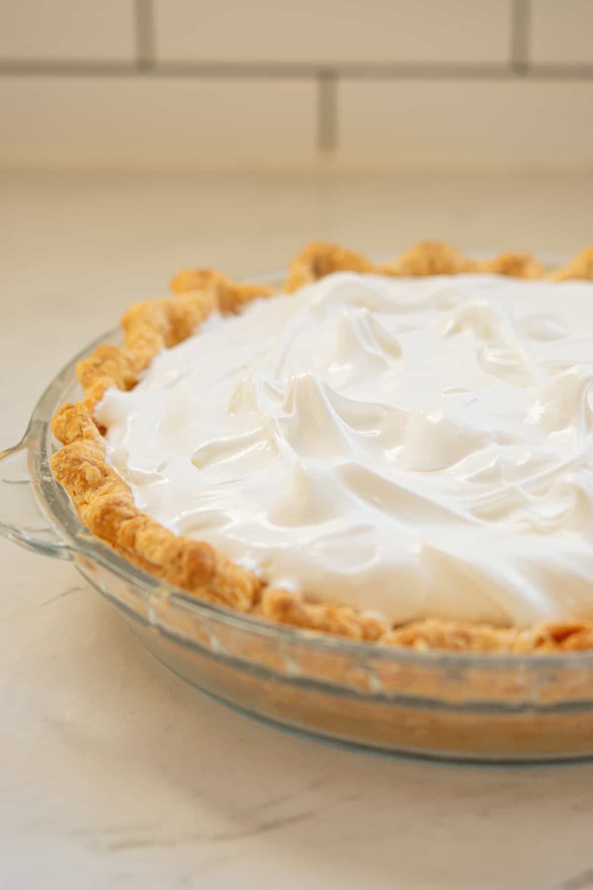 A close-up of a pie with a golden, flaky crust and a fluffy, glossy white meringue topping, displayed in a clear glass pie dish on a light-colored countertop.