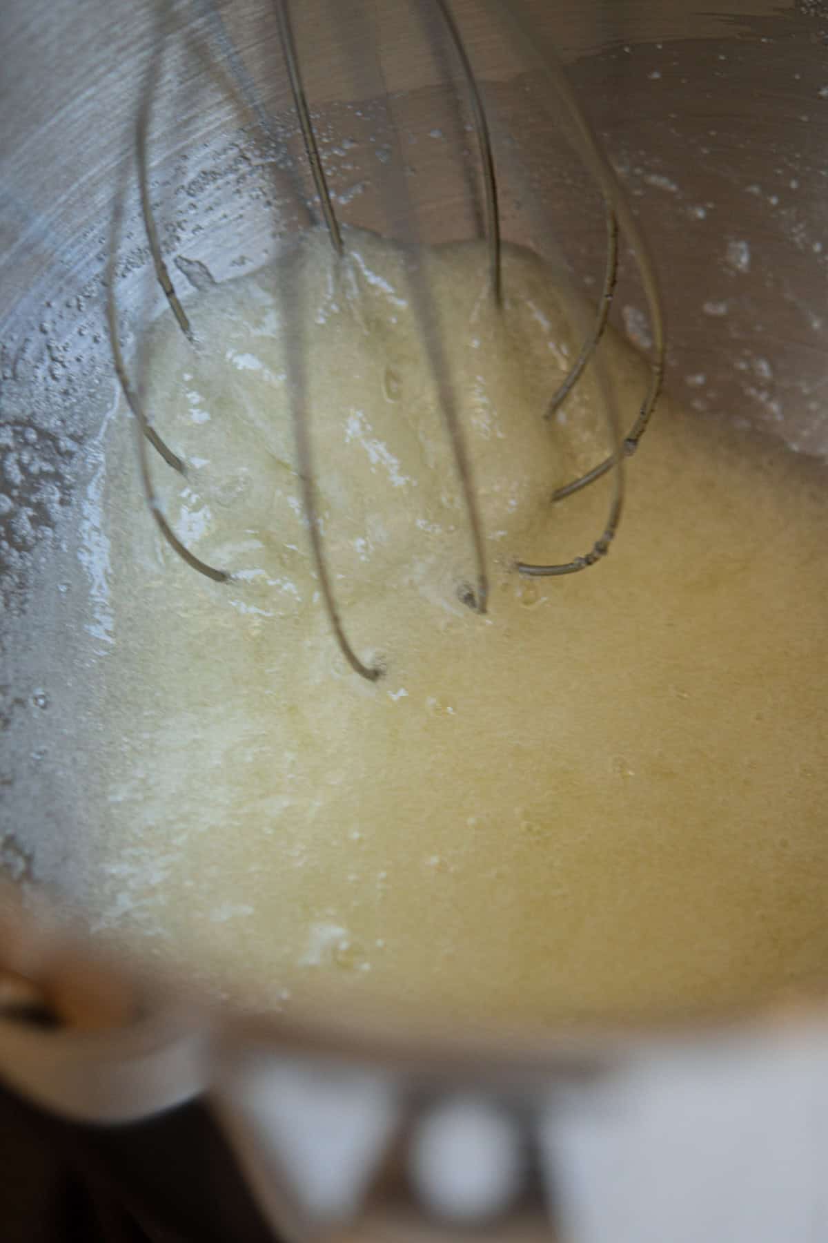 A close-up of a metal whisk mixing a pale, liquid batter inside a stainless steel mixing bowl.