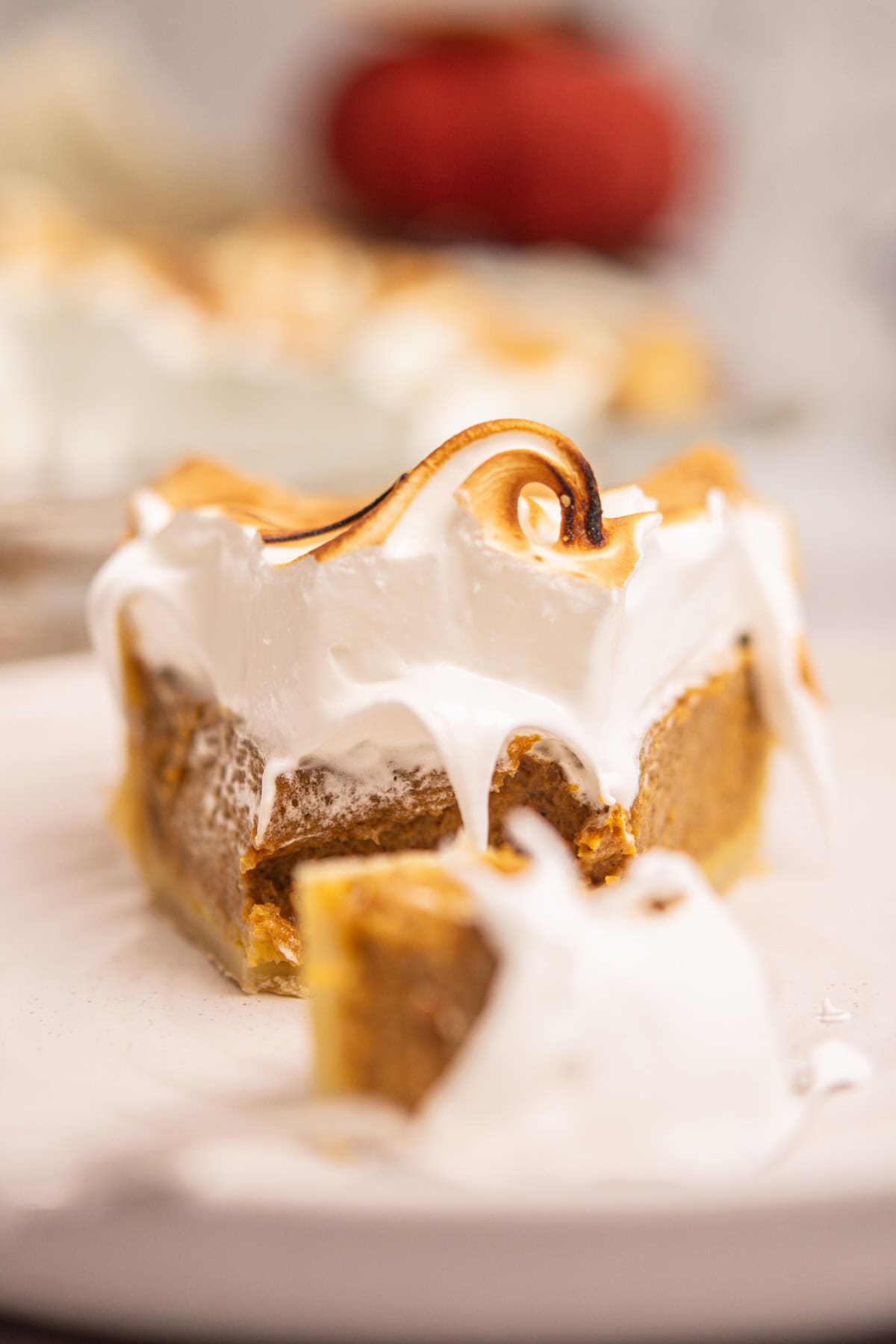 A close-up of a slice of pumpkin pie topped with toasted meringue, with a bite taken out and the creamy filling visible. The pie sits on a white plate, and the background is softly blurred.