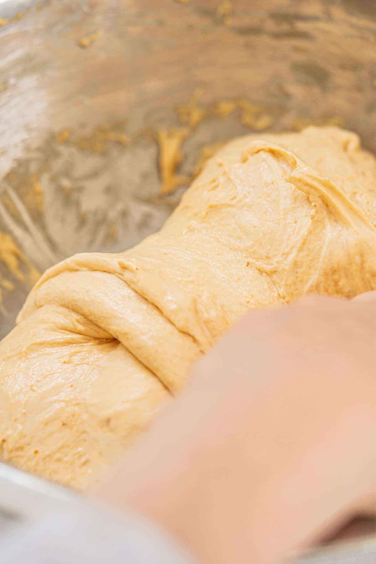 A close-up of a hand kneading soft, light brown dough in a metal mixing bowl. The dough appears smooth and elastic, with traces of flour—perfect for pairing with homemade Funfetti Ice Cream.