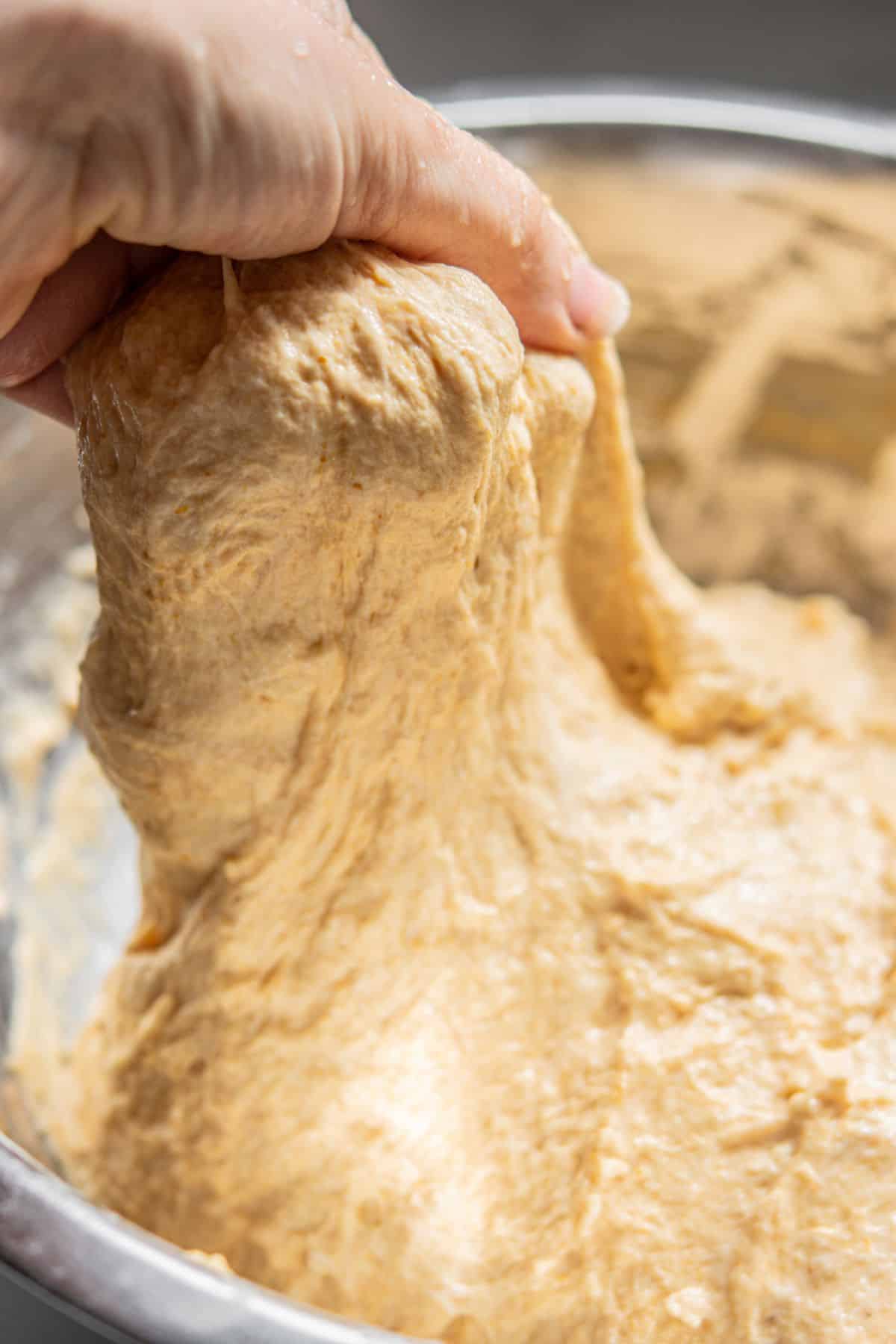 A close-up of a hand stretching sticky, risen pumpkin bread dough from a metal mixing bowl. The dough appears soft and elastic, with a slightly golden color.