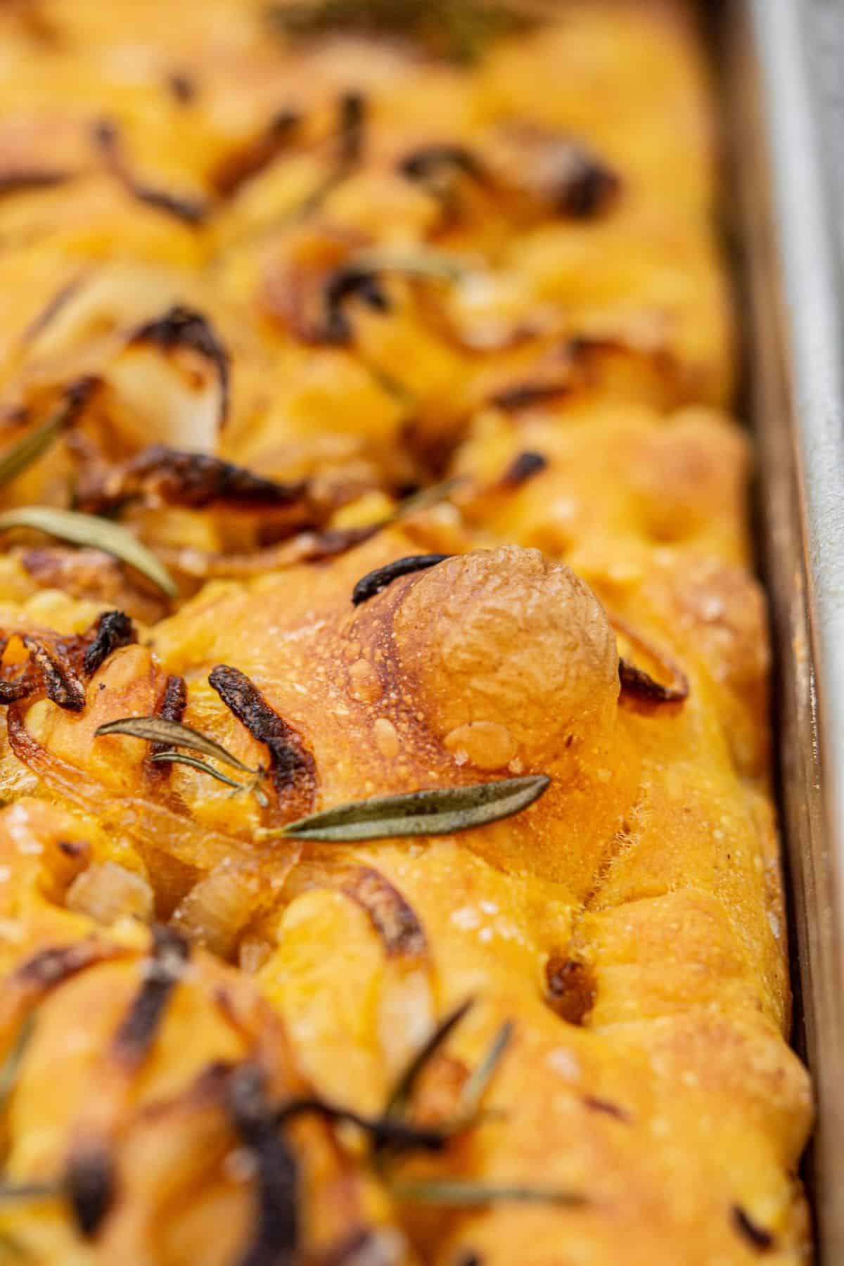 Close-up of golden-brown pumpkin focaccia bread topped with caramelized onions and fresh rosemary on a baking tray. The bread appears airy with a crisp crust and visible seasoning.