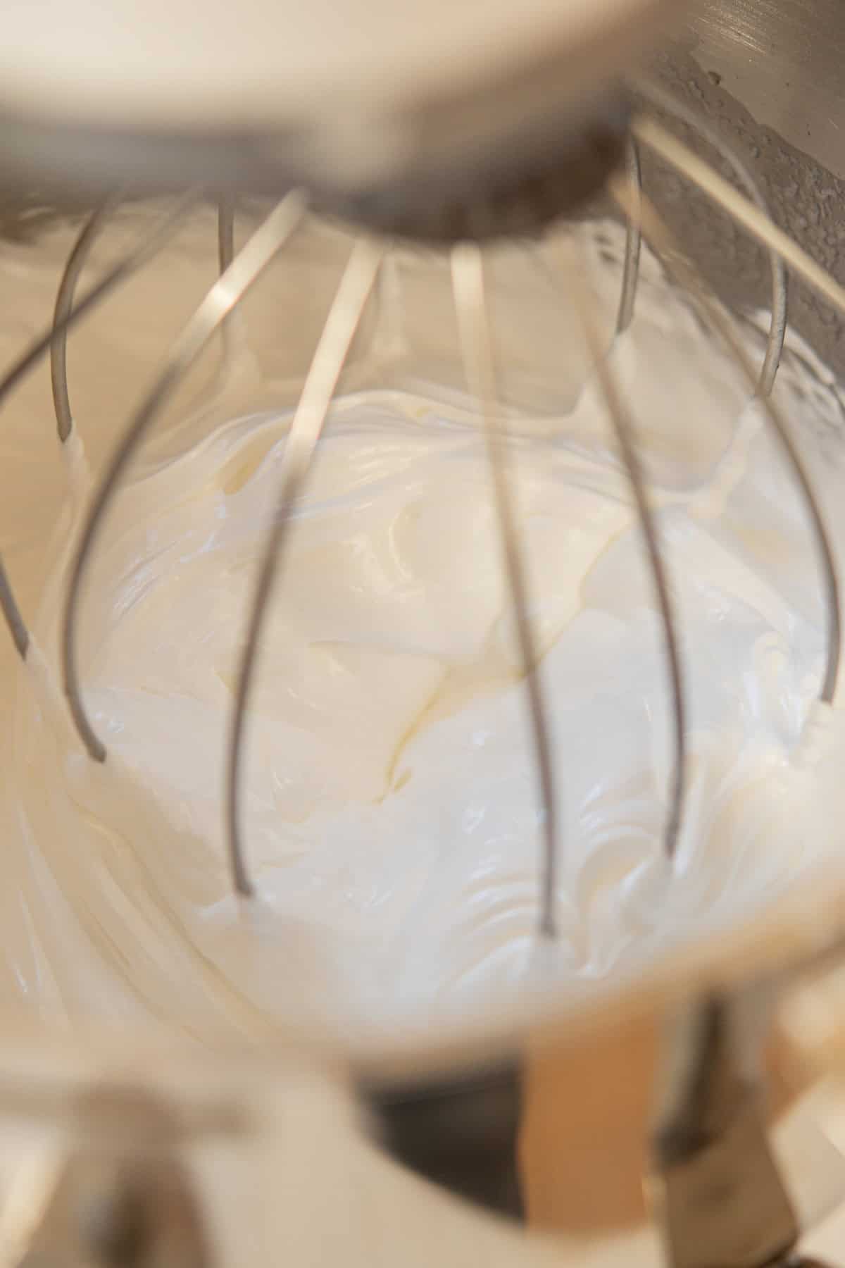 Creamy meringue being whipped in a stand mixer with a metal whisk attachment, forming stiff, glossy peaks.
