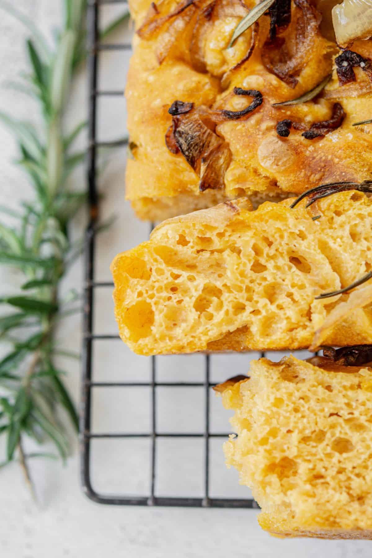 Close-up of sliced focaccia bread with a golden crust, topped with caramelized onions and rosemary, resting on a cooling rack. A sprig of fresh rosemary lies nearby on a white surface.