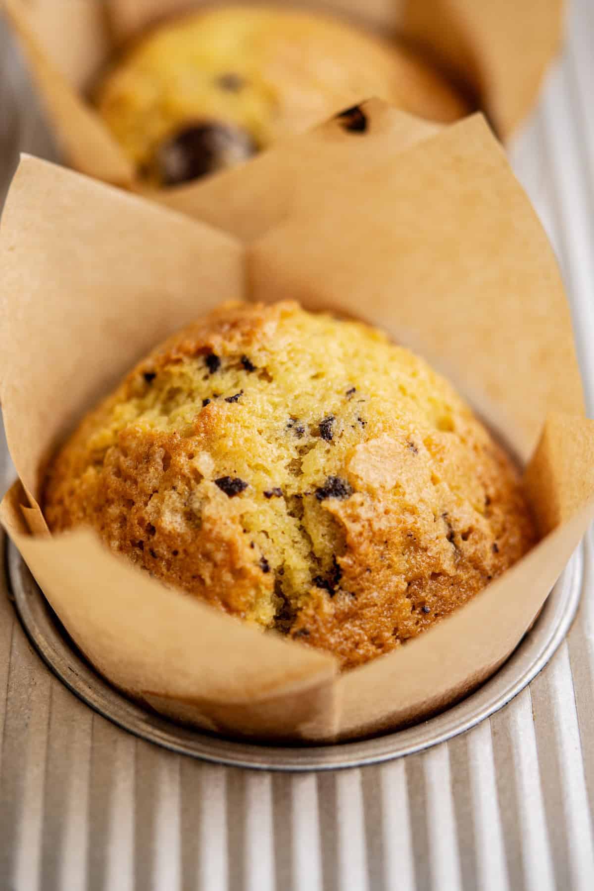 A close-up of a golden-brown orange chocolate chip muffin in a brown parchment paper wrapper, sitting in a metal muffin tray, reminiscent of the sweet speckles found in Funfetti Ice Cream.