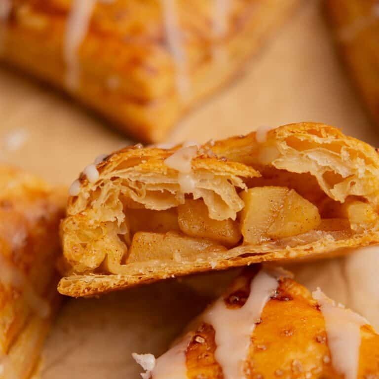 A close-up of a flaky, golden-brown pastry filled with cinnamon-spiced apple chunks, topped with a light drizzle of icing. Another whole pastry is partially visible in the background.