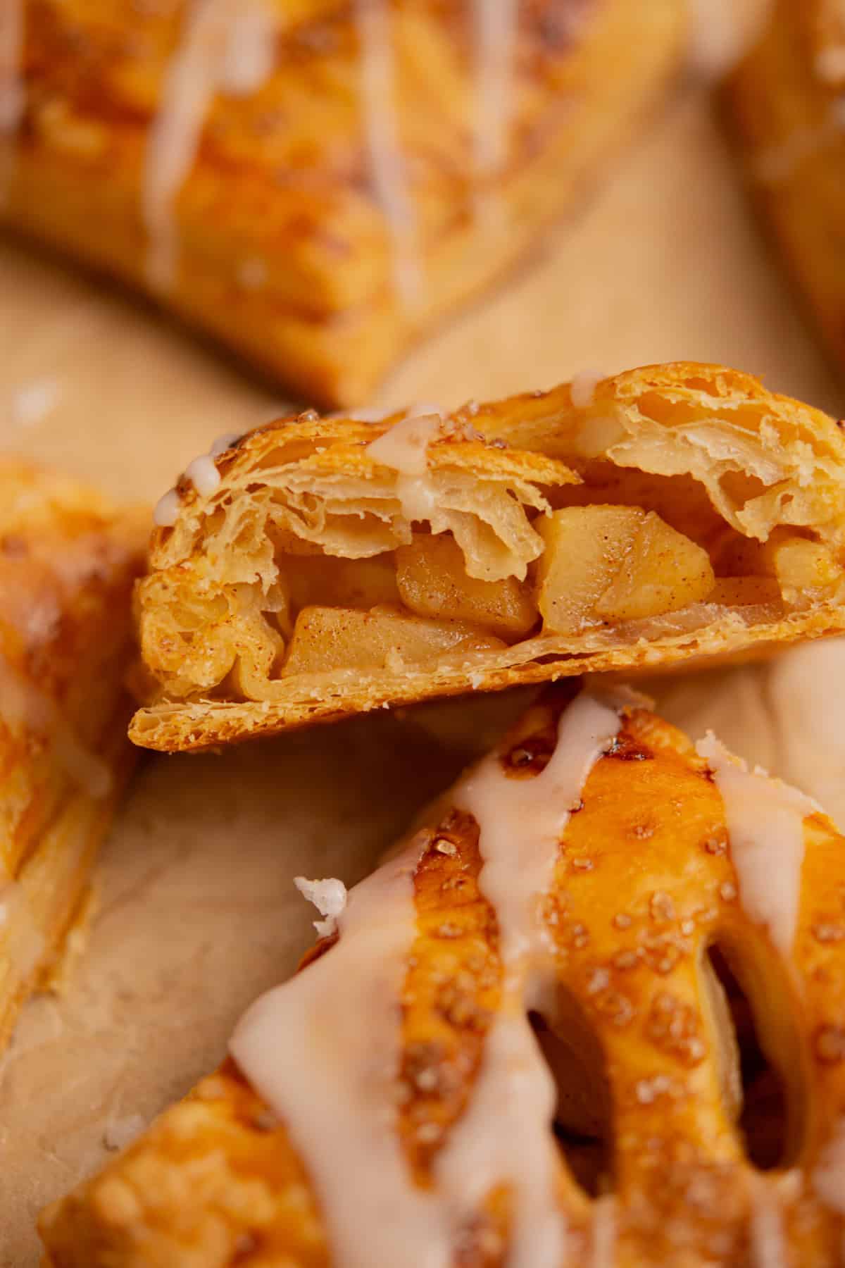 A close-up of a flaky pastry filled with spiced apple chunks, topped with a drizzle of white icing. The pastry is golden brown and shown on parchment paper.