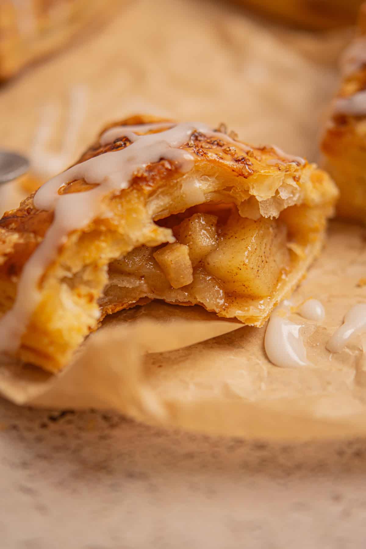 A close-up of Apple Hand Pies with Puff Pastry, filled with diced apples and cinnamon, topped with white icing and resting on brown parchment paper. One end is bitten, revealing the sweet apple filling inside.