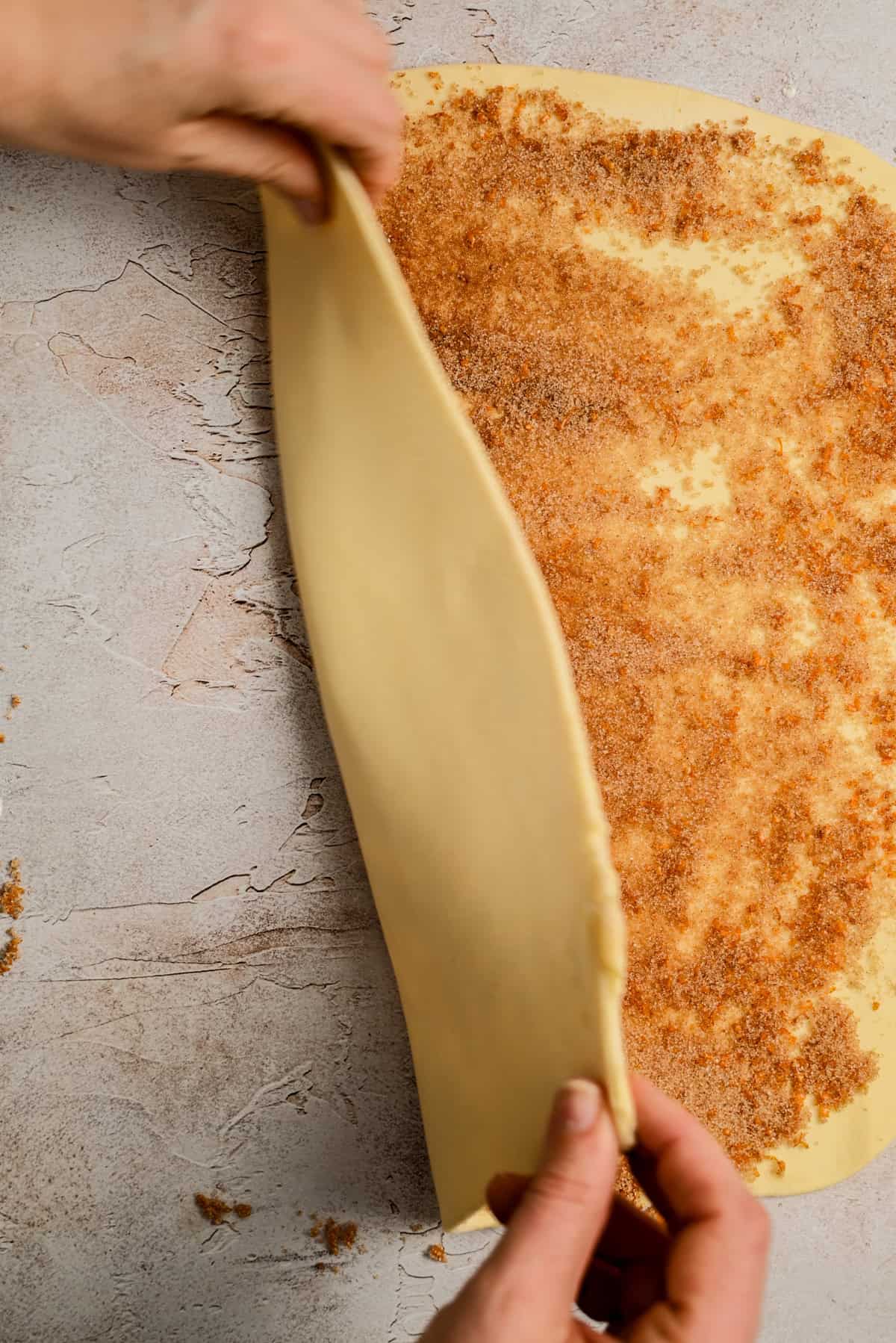 Hands begin to roll up a sheet of dough topped with a brown sugar and cinnamon mixture on a light textured surface, preparing to make cinnamon rolls.