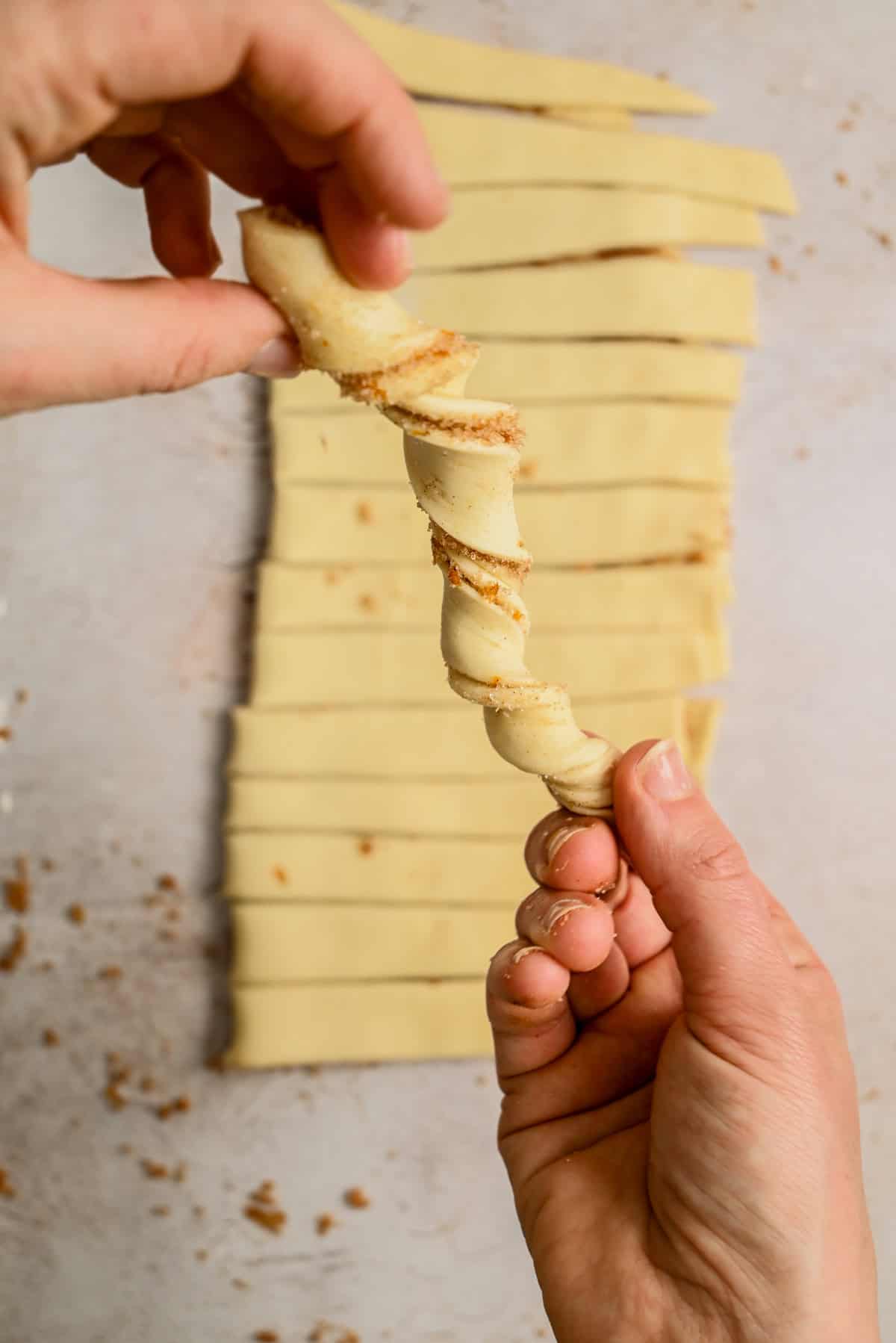 Two hands twist a strip of dough coated with cinnamon sugar, with more dough strips and crumbs visible on the surface below.