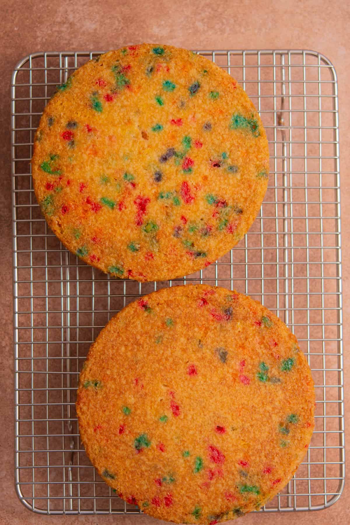 Two round funfetti cake layers with colorful sprinkles are cooling on a metal wire rack over a brown surface.