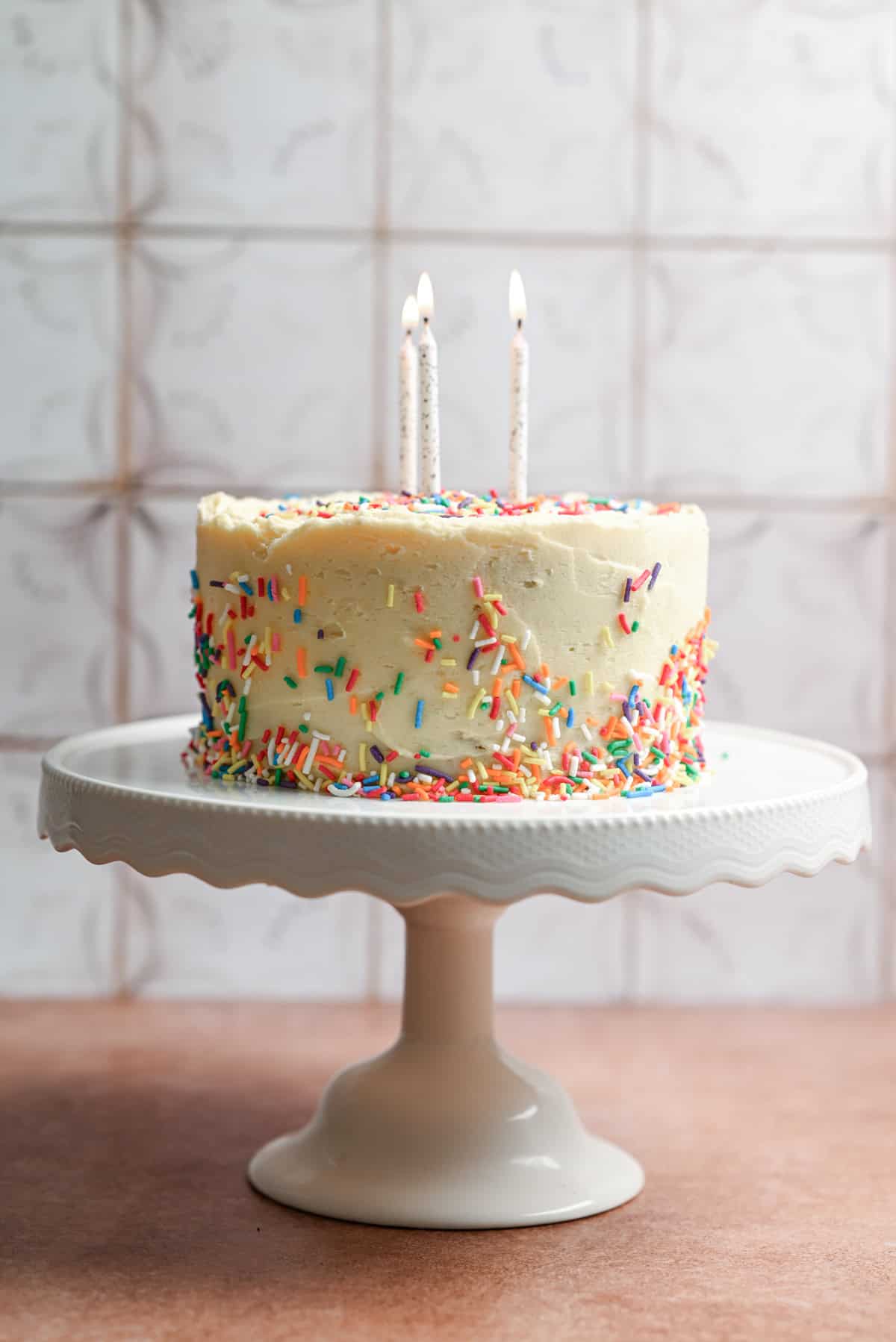 A frosted layer cake decorated with colorful sprinkles and topped with two lit candles sits on a white cake stand, while Puff Pastry Cinnamon Twists add a delightful touch against the tiled background.