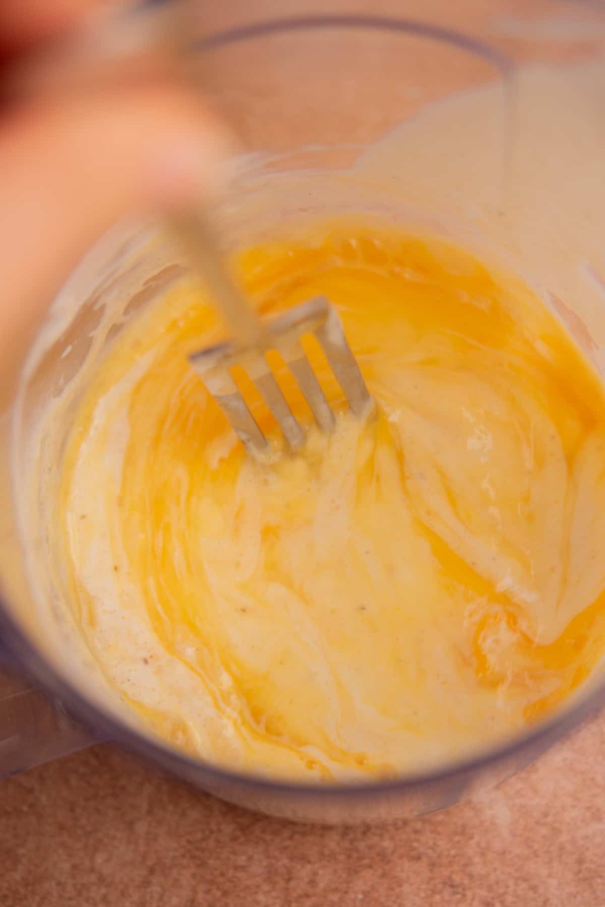 A close-up of a hand mixing eggs and batter with a fork in a glass bowl, creating a swirled yellow and creamy mixture.
