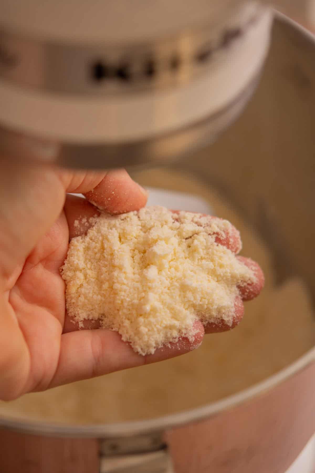A close-up of a hand holding a small pile of crumbly, pale yellow dough mixture above a metal mixing bowl, with a stand mixer in the background.