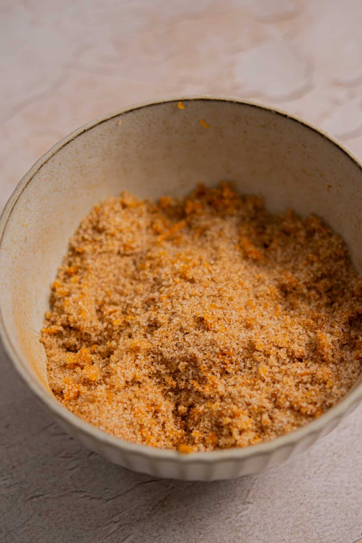 A ceramic bowl filled with a crumbly mixture of brown sugar and orange zest, sitting on a light-colored textured surface.