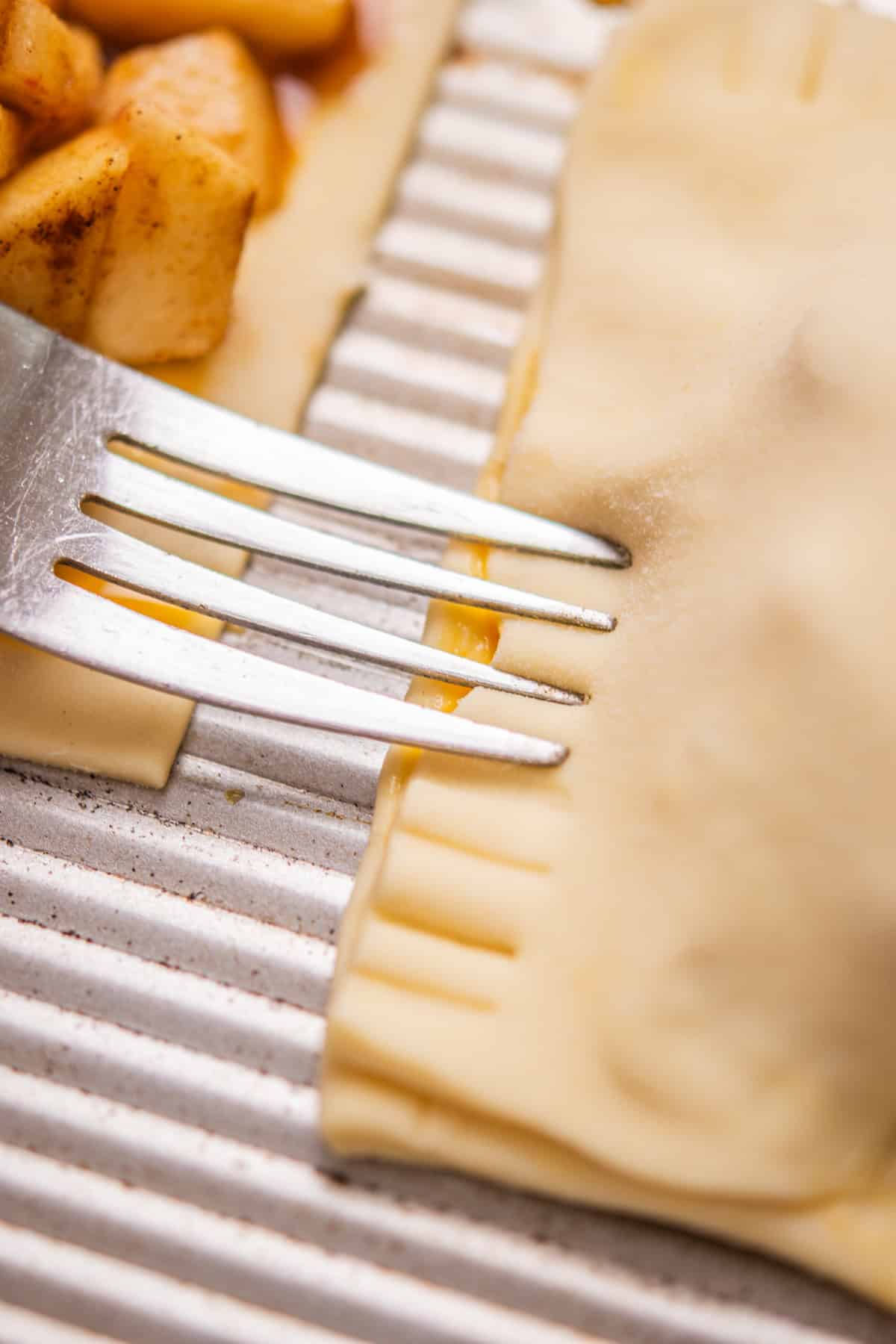 A close-up of a fork pressing the edges of a pastry pocket to seal it, with some apple filling visible in the background on a ridged baking sheet.