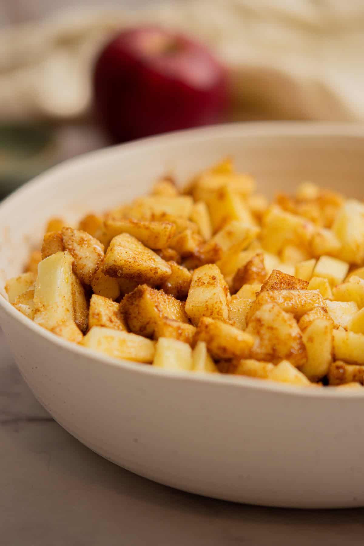 A close-up of a bowl filled with chopped apples coated in cinnamon, with a blurred red apple in the background.