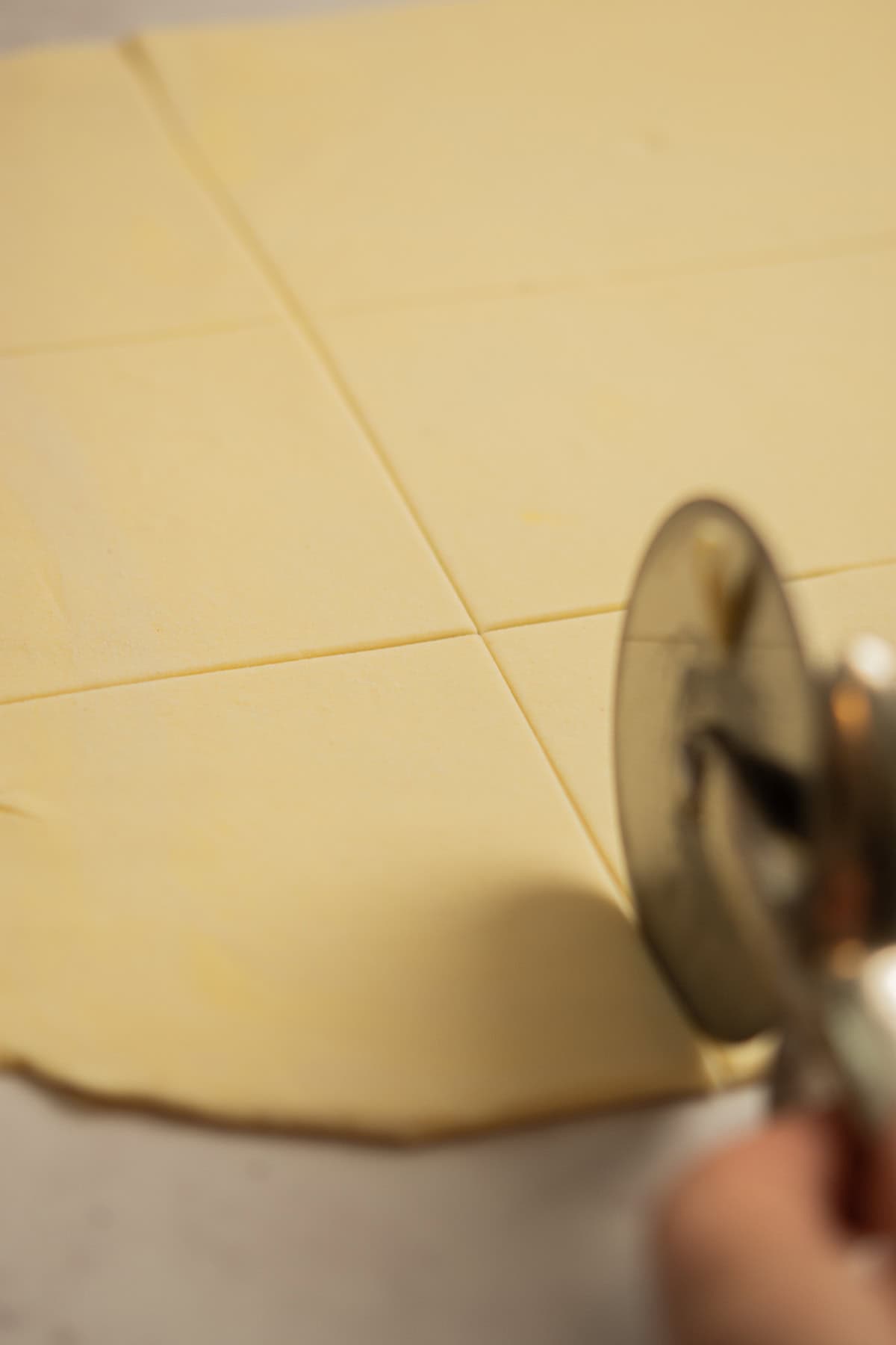 A hand uses a pizza cutter to cut a sheet of dough into squares on a flat surface.