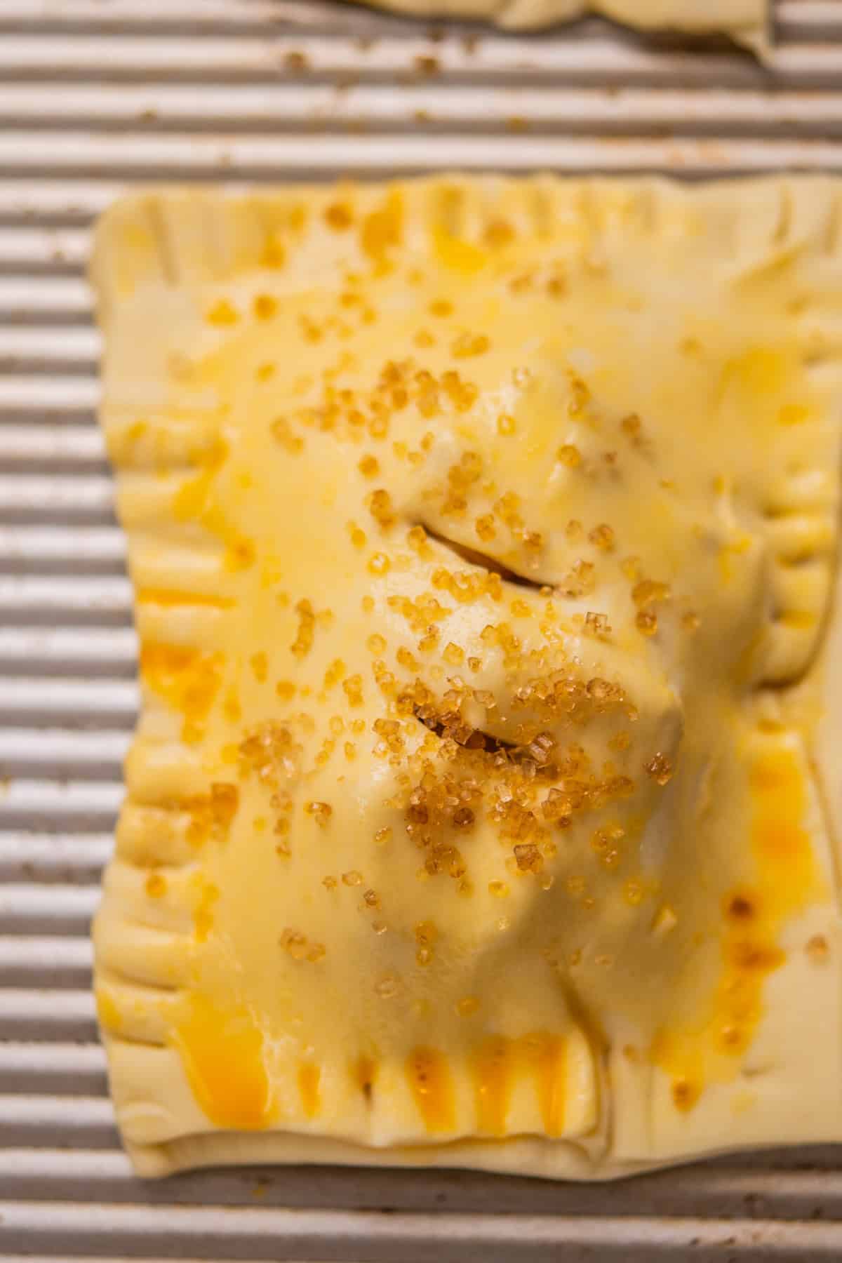 A close-up of an unbaked pastry pocket on a ridged baking sheet, brushed with egg wash and sprinkled with coarse sugar crystals, with forked edges and small slits on top.