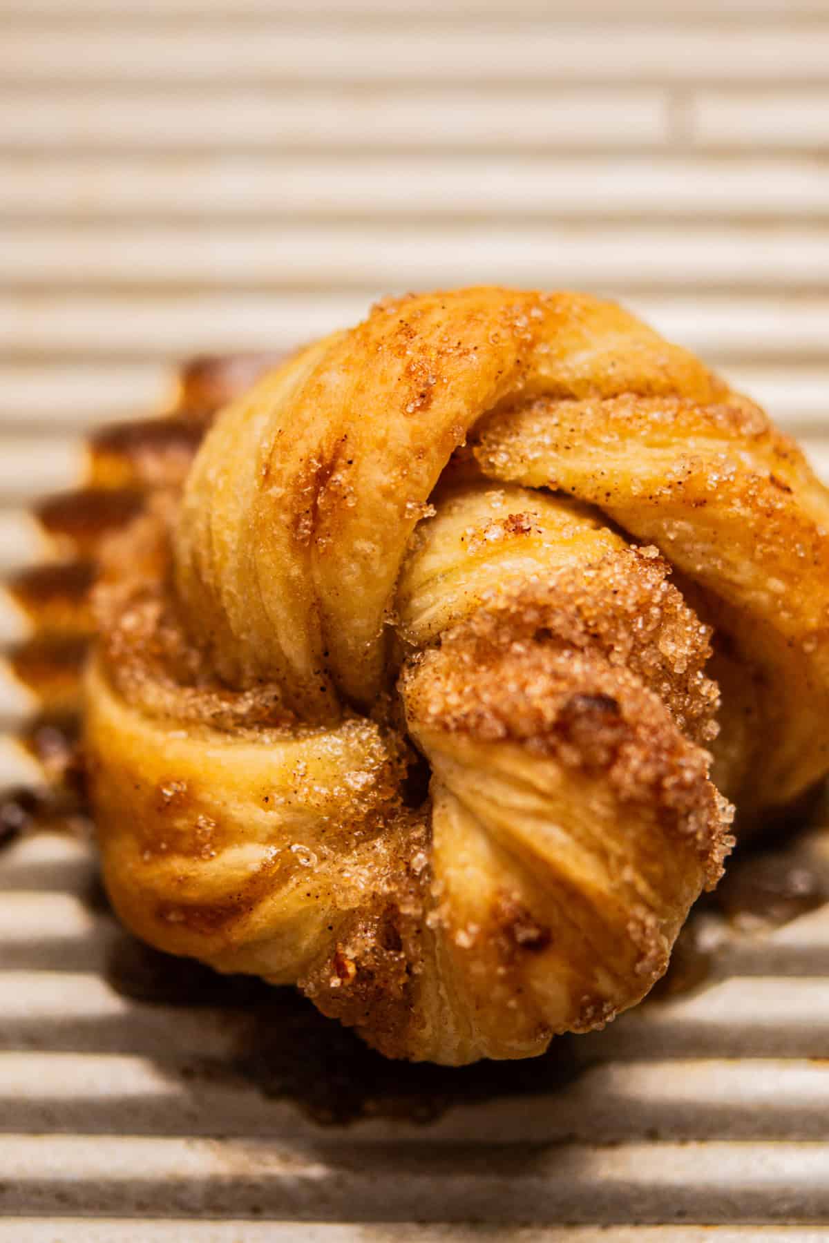 A close-up of a golden-brown, twisted pastry dusted with sugar and cinnamon, resting on a ridged, light-colored baking tray.