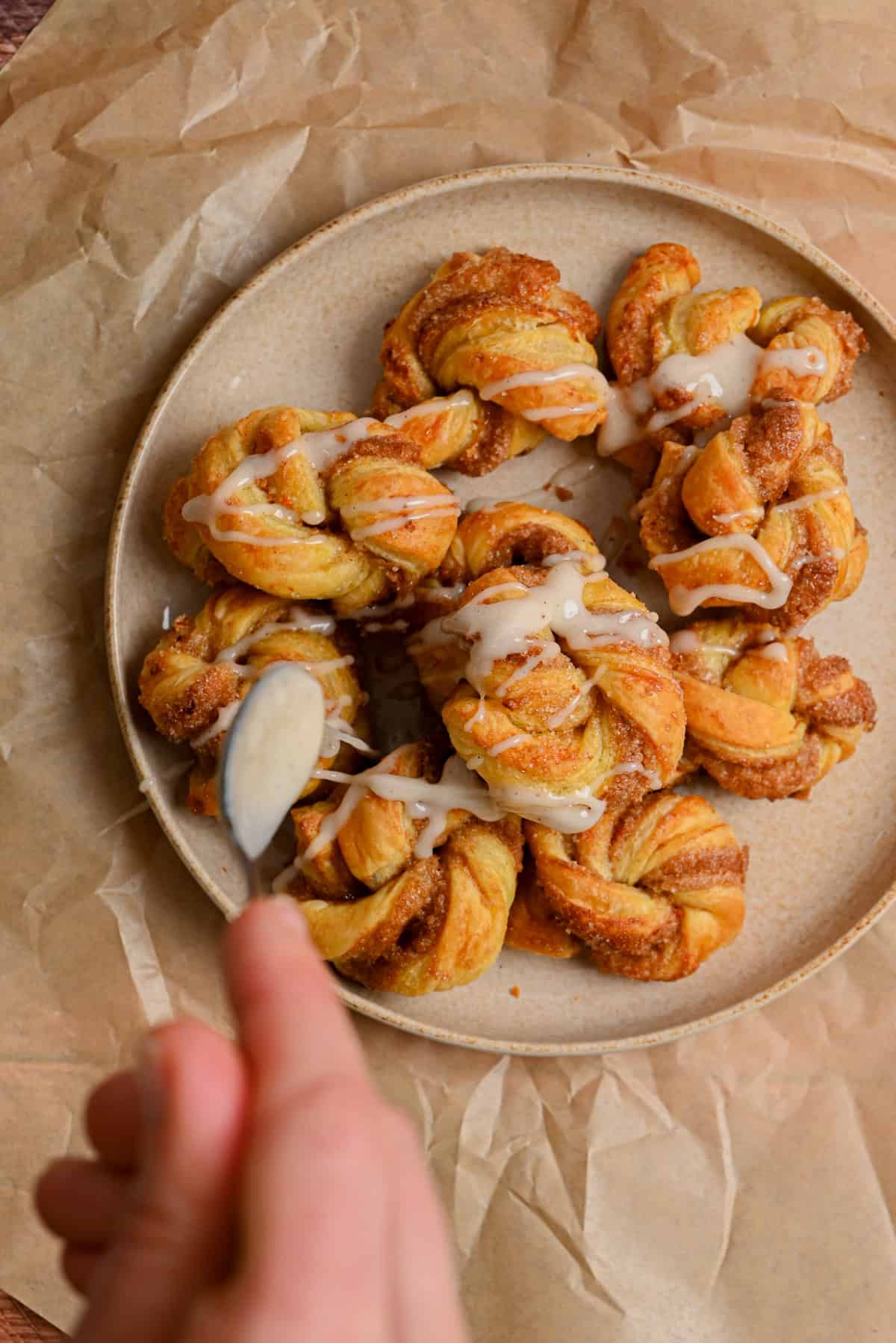 A hand drizzles white icing over a plate of twisted, golden-brown pastries on parchment paper. The pastries appear freshly baked and are displayed in a pleasing arrangement.