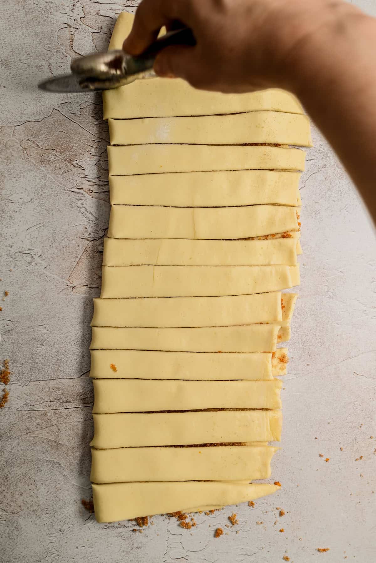 A hand uses a pastry wheel to cut a sheet of dough into even strips on a light-colored surface.