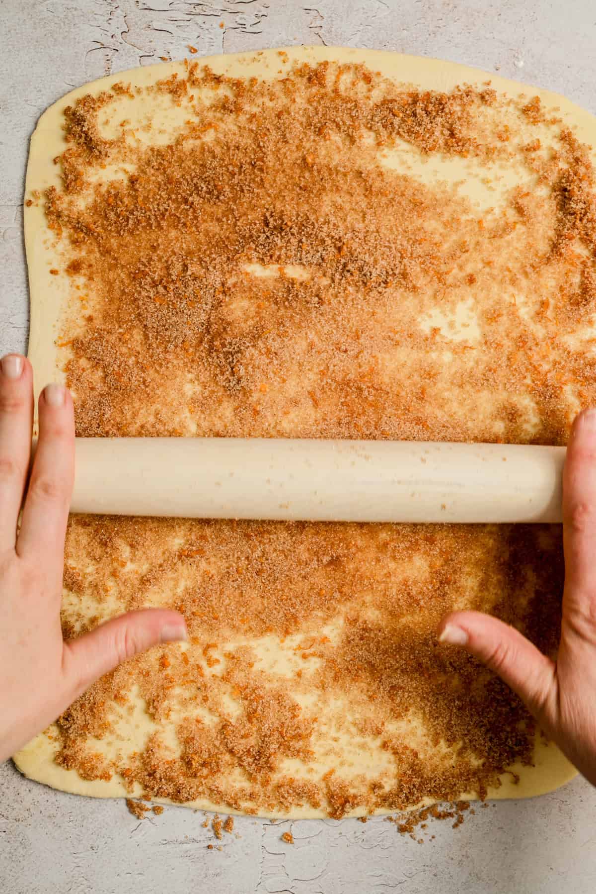 Hands rolling up a sheet of dough topped with a mixture of brown sugar and cinnamon on a lightly floured surface, preparing to make cinnamon rolls.