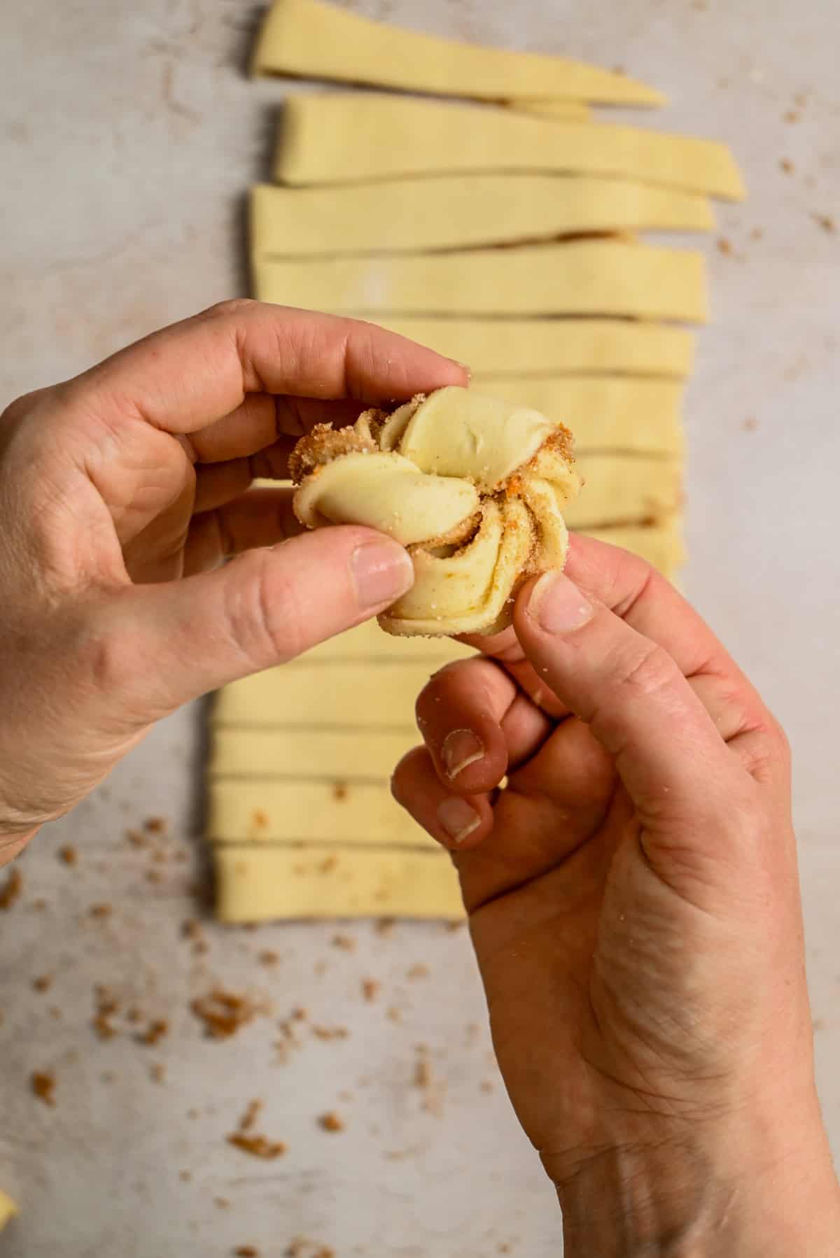 Two hands shape a piece of dough filled with cinnamon and sugar, with several rectangular dough strips and crumbs visible in the background on a light surface.