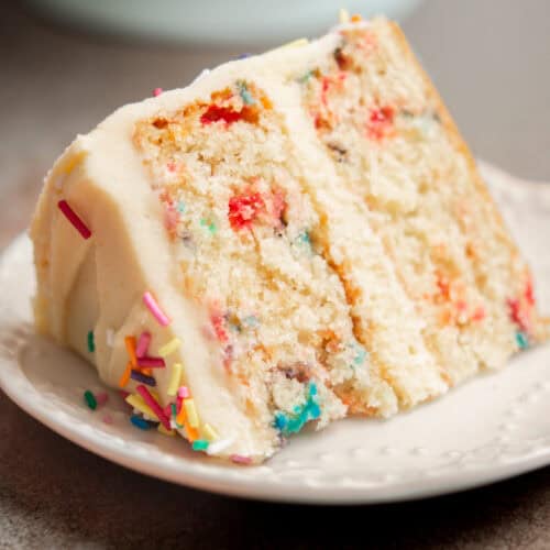 A slice of funfetti cake with white frosting and colorful sprinkles sits on a white plate, next to flaky Puff Pastry Cinnamon Twists. The cake has two layers with rainbow sprinkles baked inside.