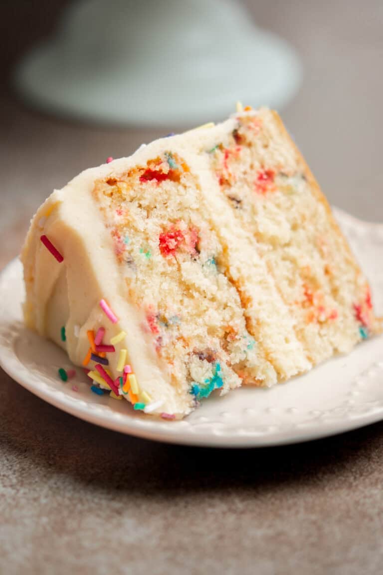 A slice of funfetti cake with white frosting and colorful sprinkles sits on a white plate, next to flaky Puff Pastry Cinnamon Twists. The cake has two layers with rainbow sprinkles baked inside.